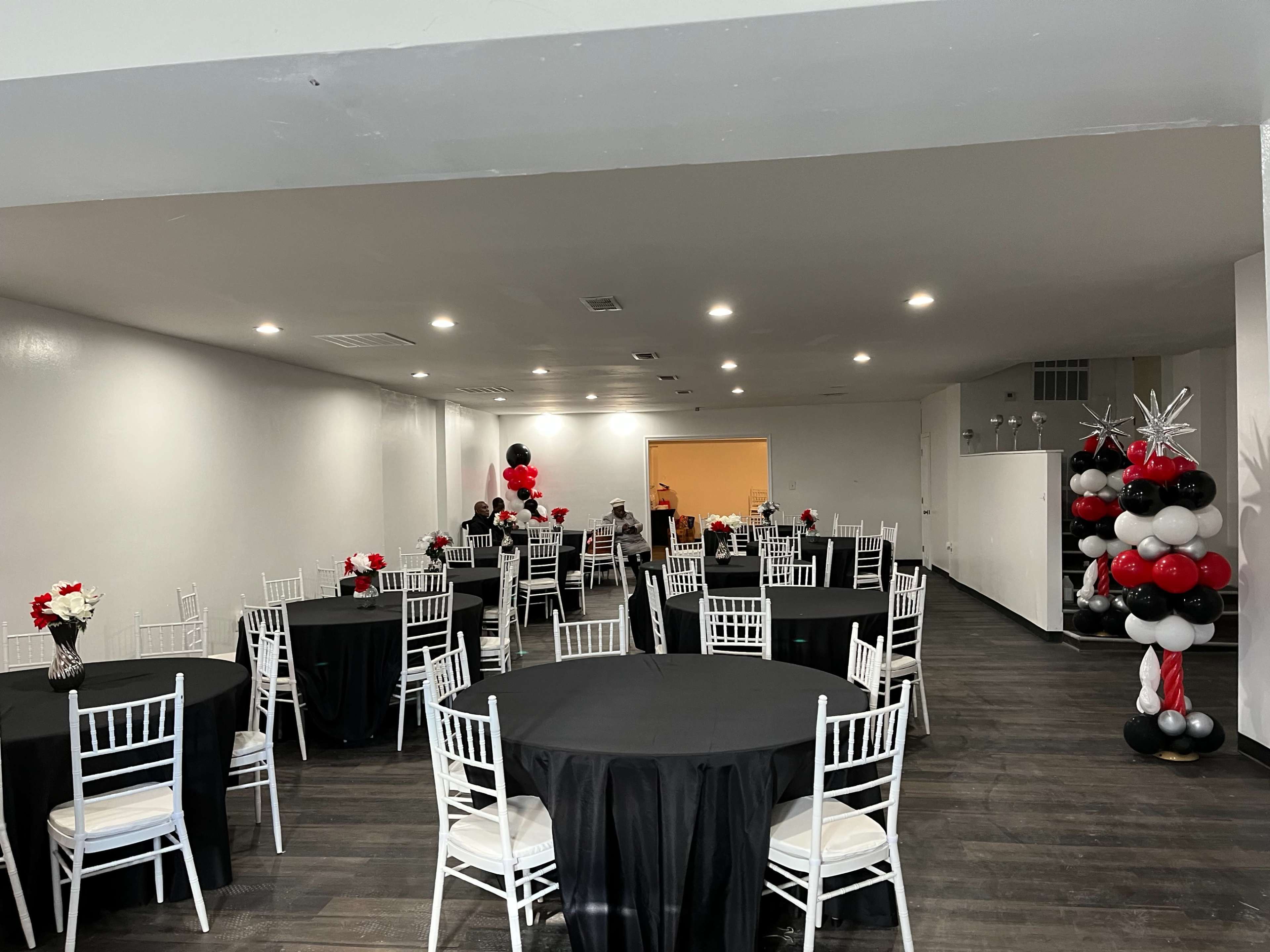 The image shows an indoor event space with round black tables and white chairs arranged for a gathering, decorated with red and white floral centerpieces and a balloon display in the corner.