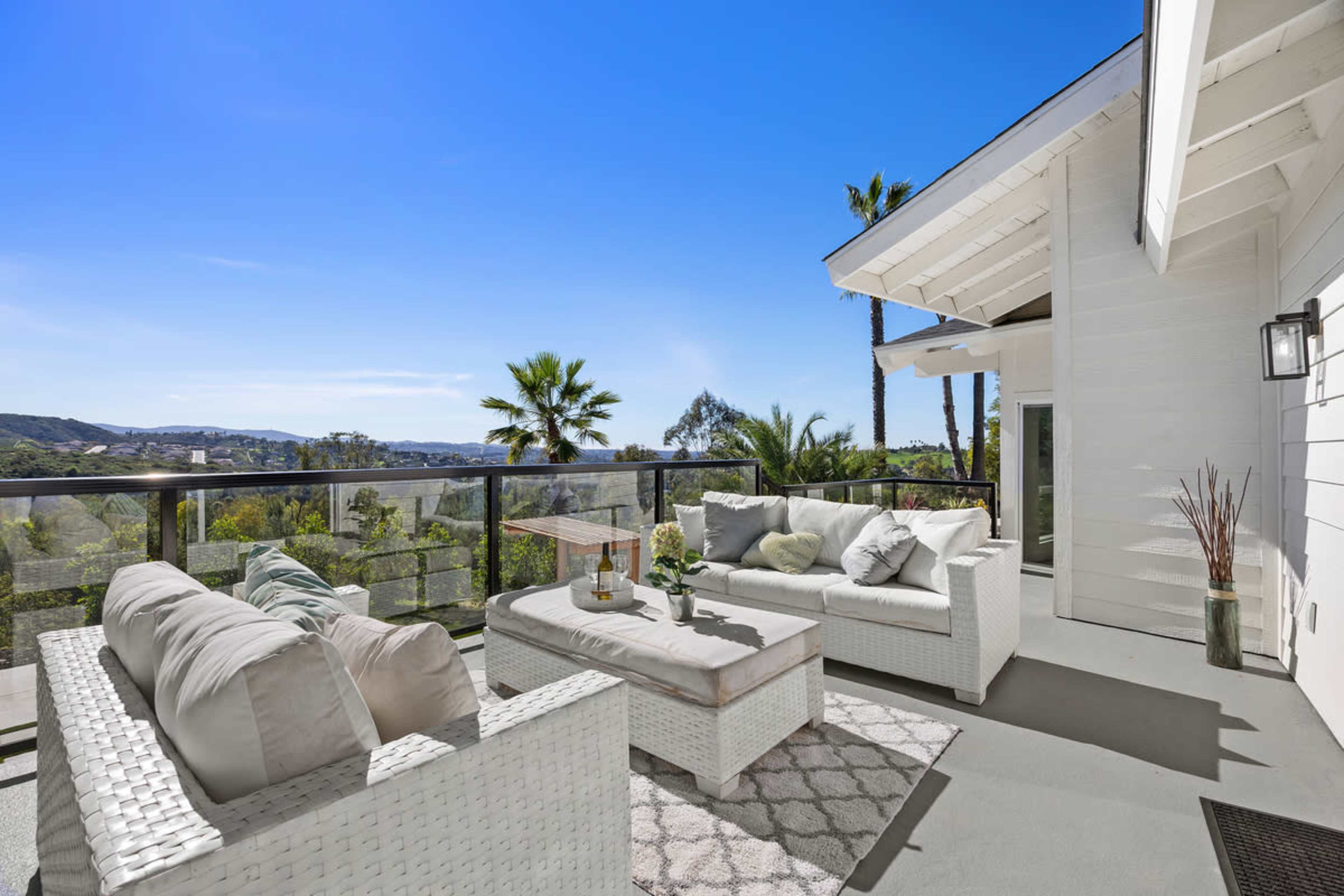 The image shows a spacious outdoor patio with white wicker furniture, a coffee table, and a clear view of palm trees and hills under a bright blue sky.