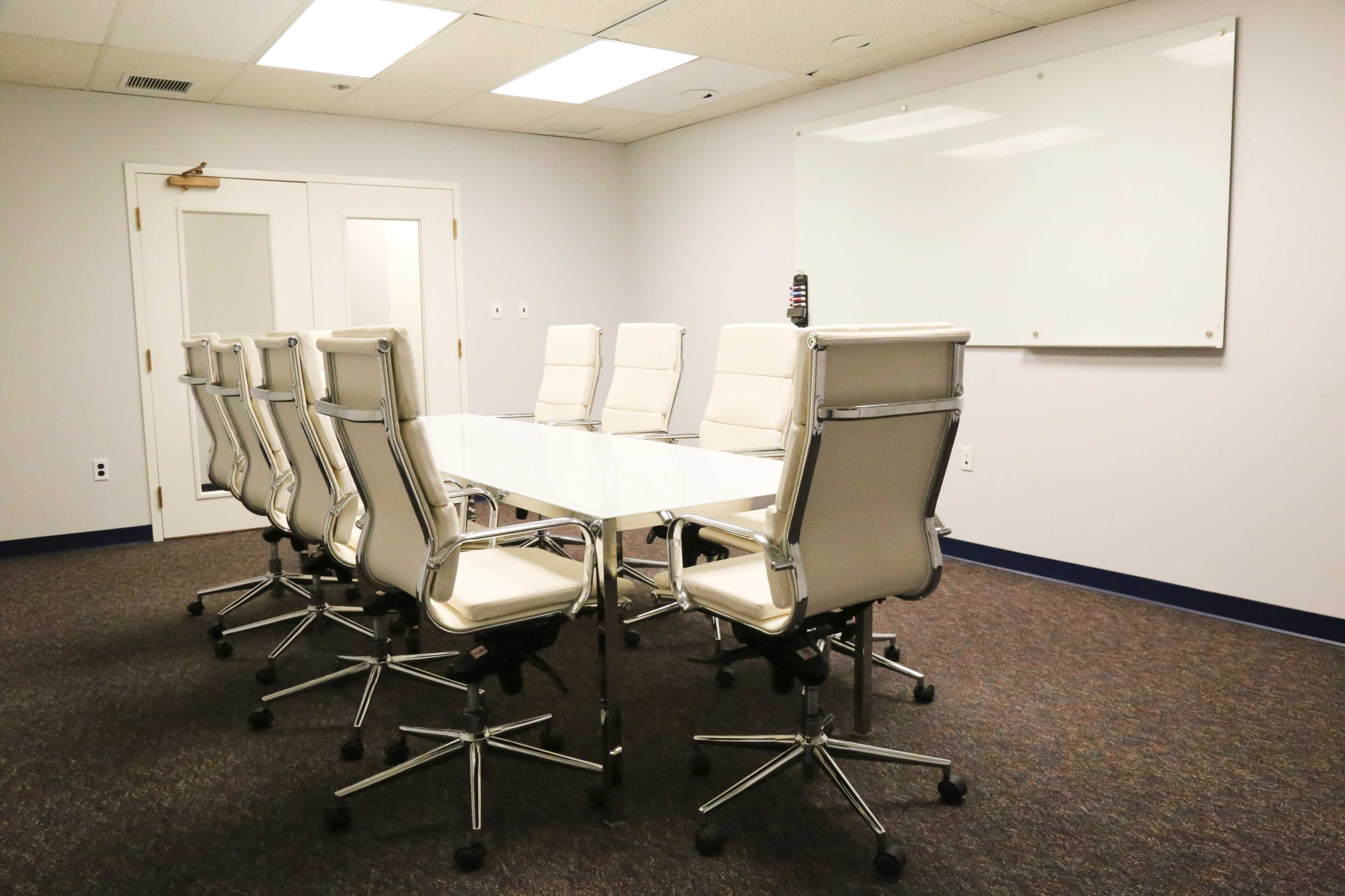 A modern conference room with a long glass table surrounded by eight white ergonomic chairs and a whiteboard mounted on the wall.