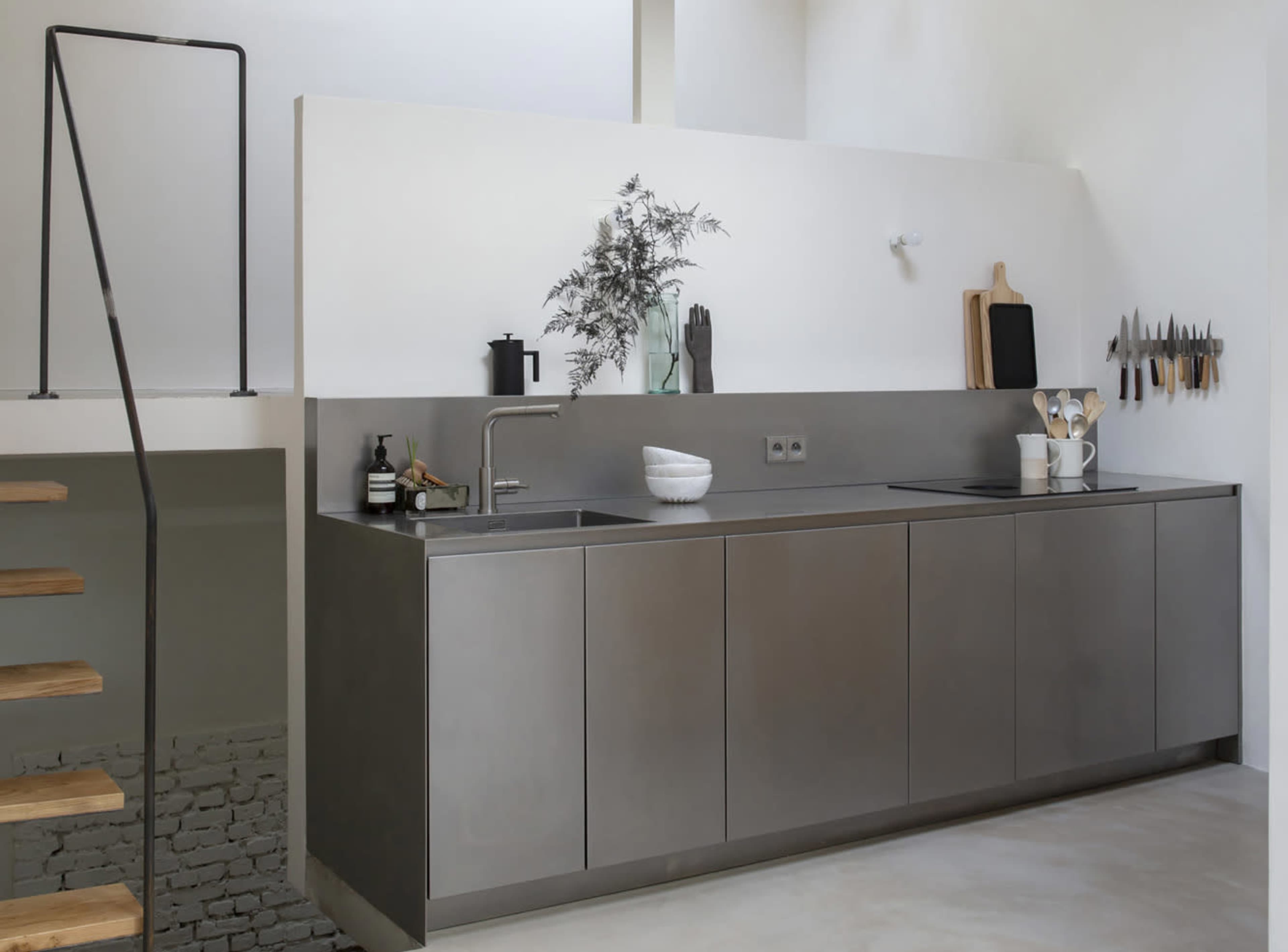 The image shows a modern kitchen with a stainless steel countertop and cabinetry, featuring minimal decor and a staircase in the background.