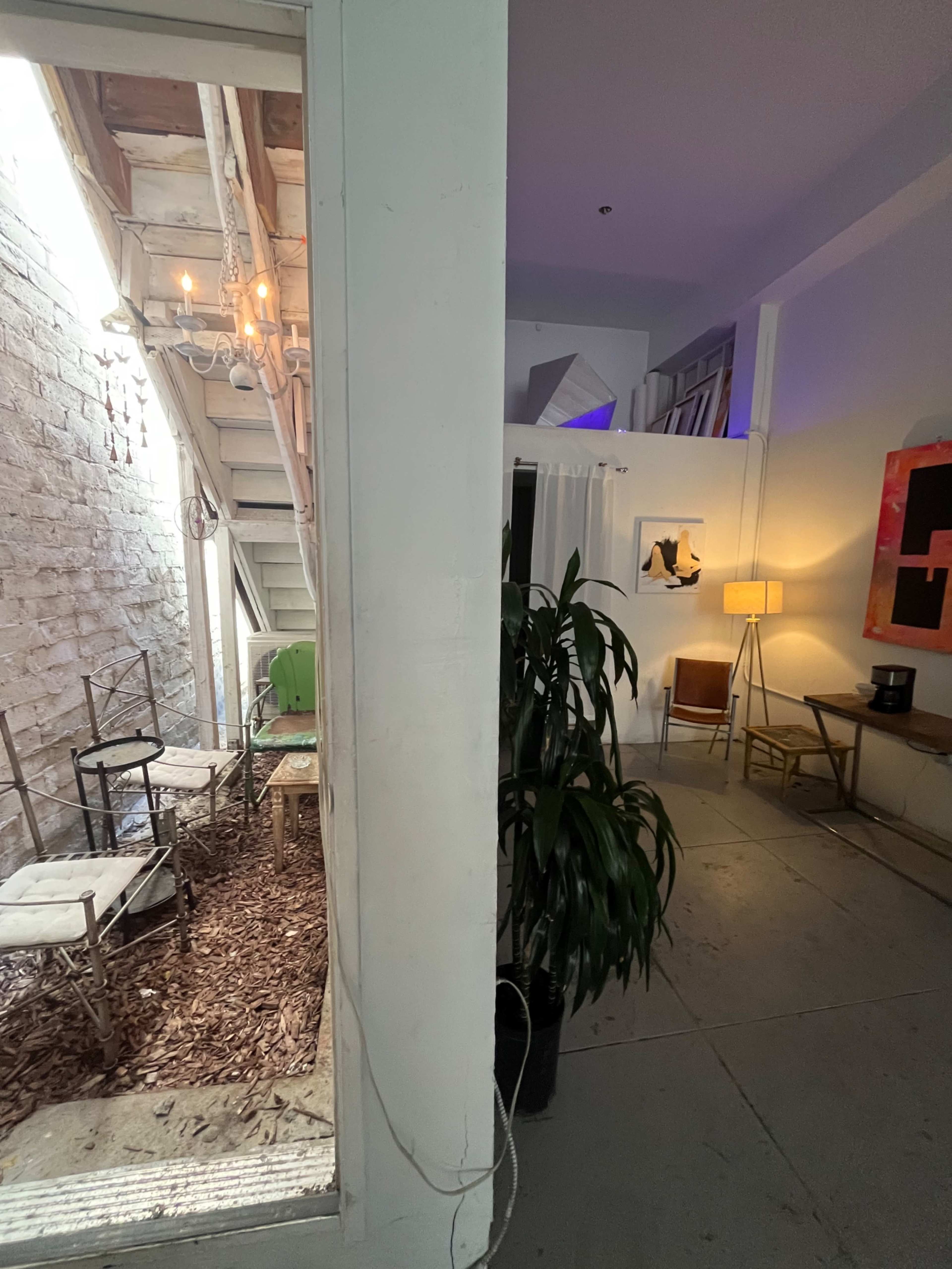 A split view of an indoor space with a small outdoor area featuring chairs and a staircase, while the other side contains a lit room with a lamp, art on the wall, and a coffee maker.