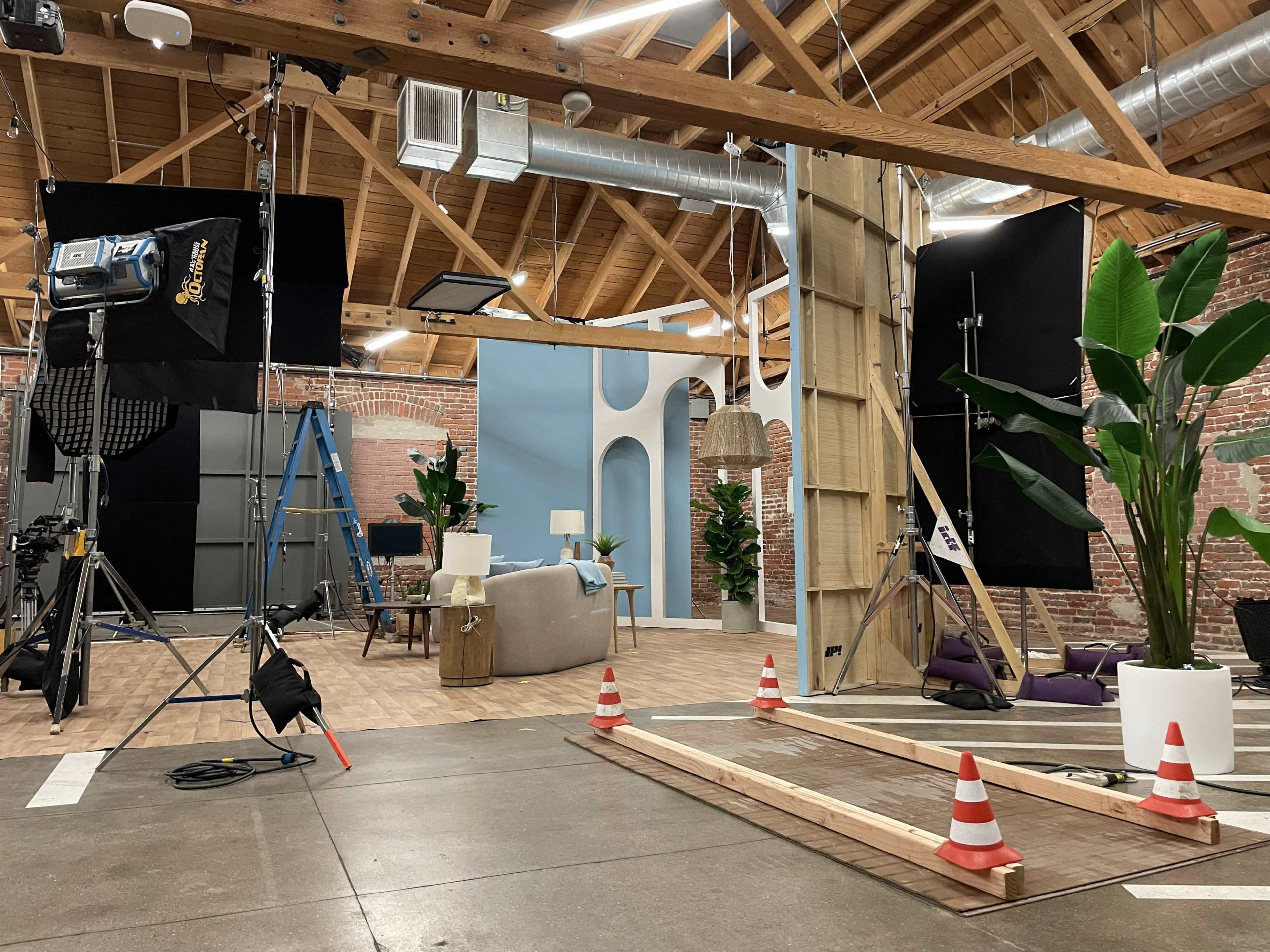 A production studio with various lighting equipment, set pieces, and construction materials, surrounded by potted plants and caution cones.