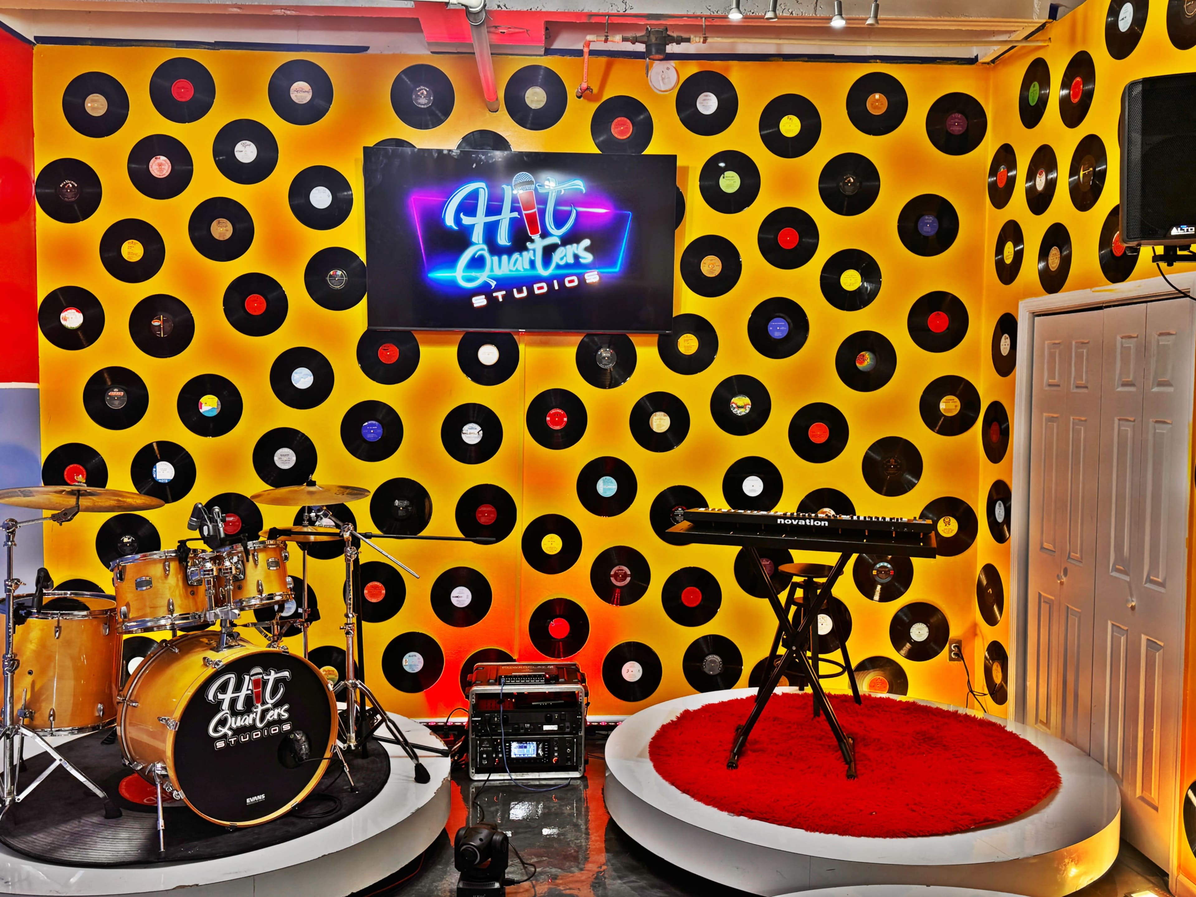 The image showcases a music studio setting with a drum set and keyboard on a vibrant wall decorated with vinyl records and a neon sign reading "Hit Quarters Studio."