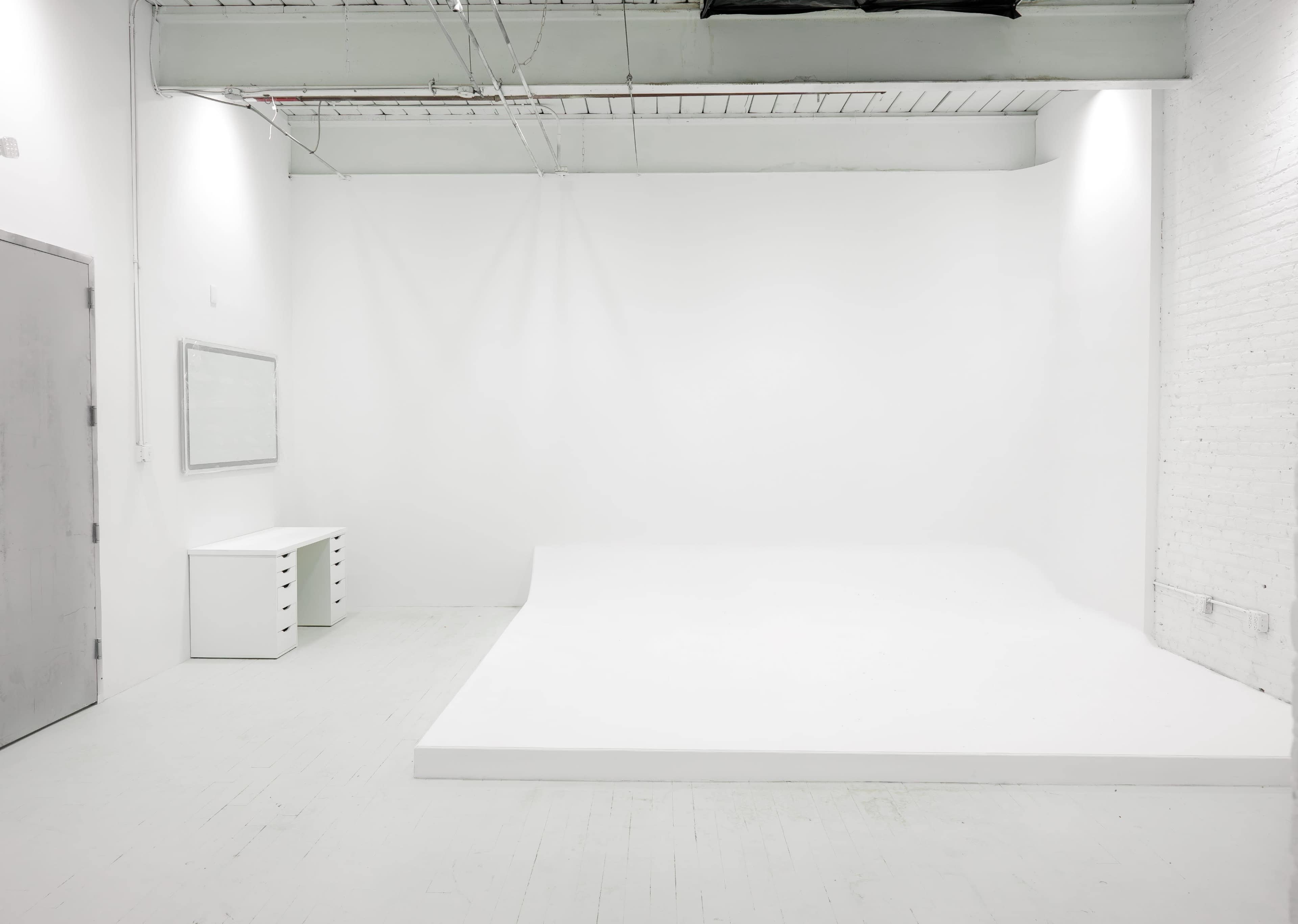 A minimalistic white studio space with a large seamless backdrop and a small set of drawers against one wall.
