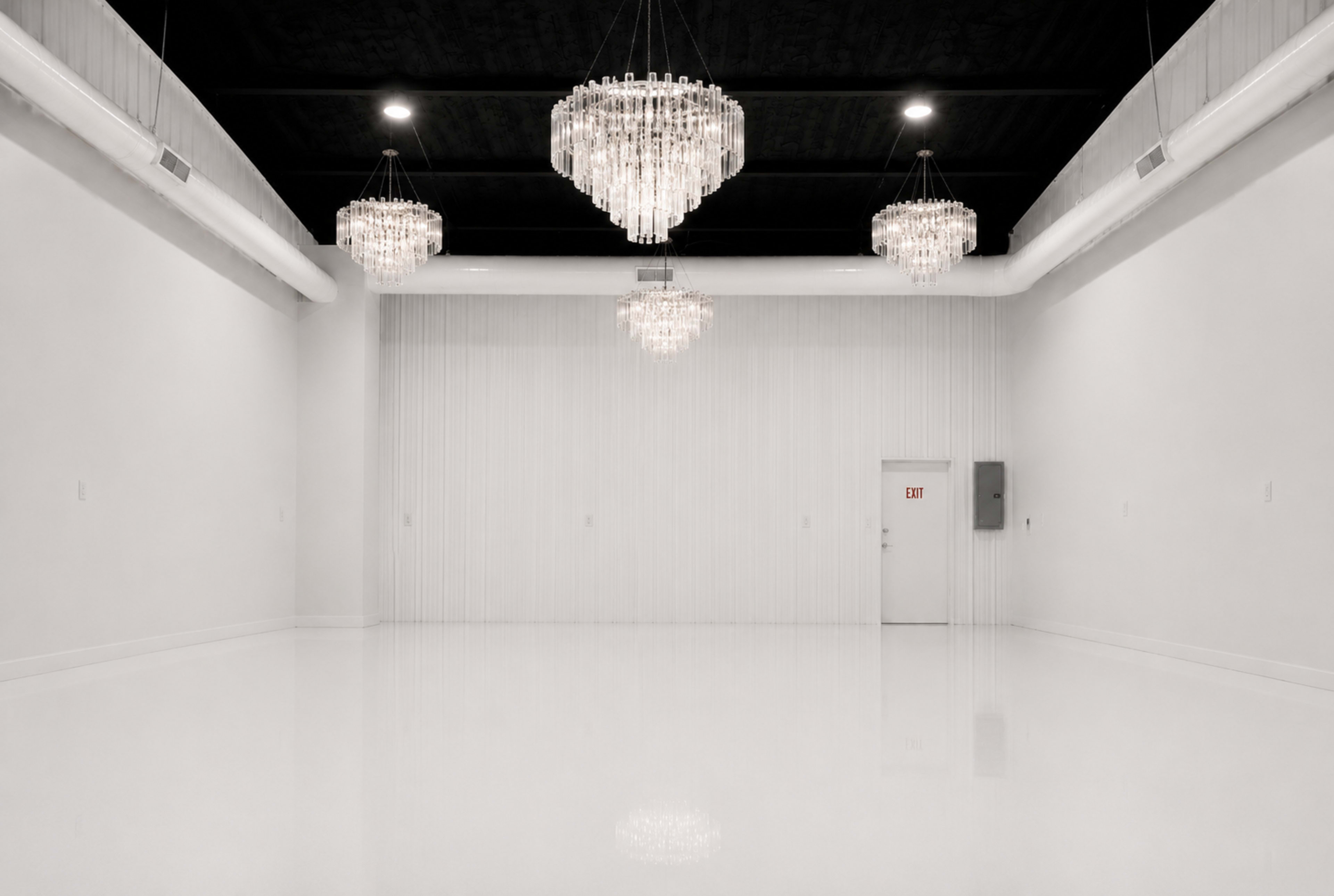 The image features a large, minimalistic white room with several crystal chandeliers hanging from the ceiling and a single door on one wall.