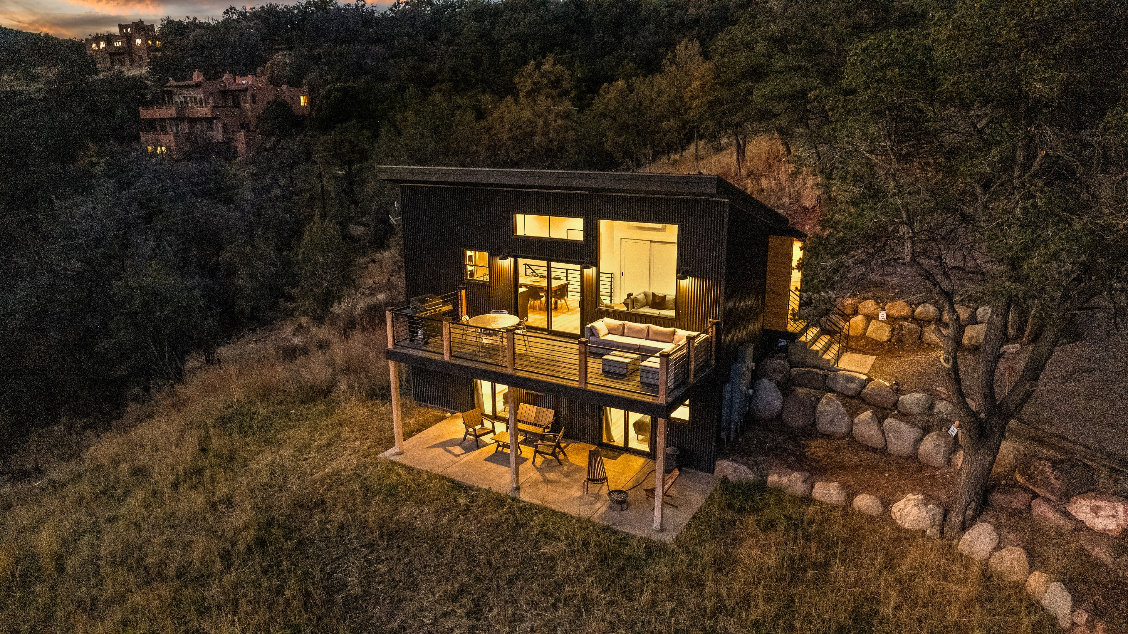 The image shows a modern black house with large windows, situated on a hillside surrounded by trees and rocks, illuminated by warm interior lights.