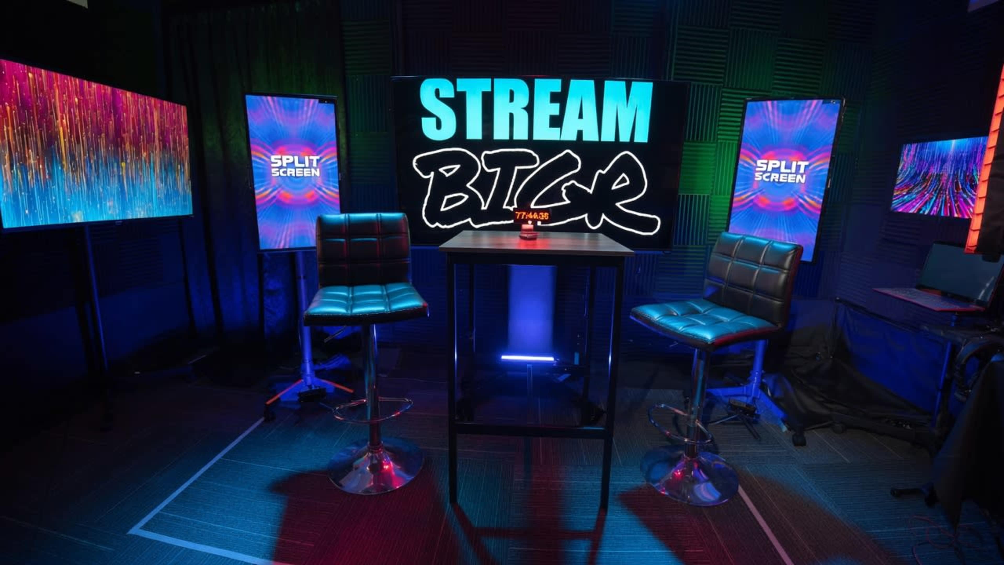 A modern video streaming setup features two high stools around a central table, with vibrant screens displaying colorful graphics and the word "STREAM" in bold letters in the background.