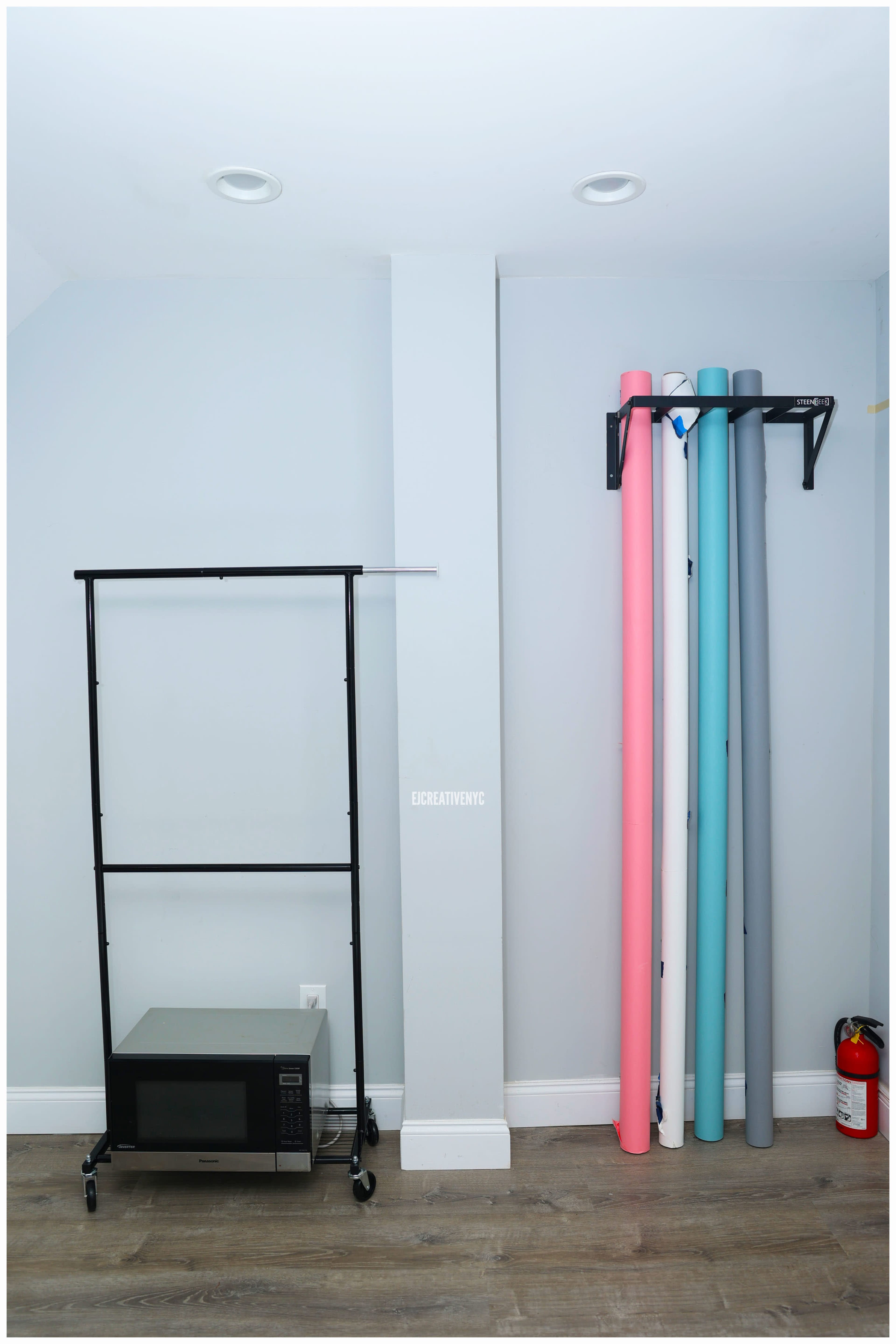 A room with a black clothes rack, a microwave on a stand, and rolls of colored paper leaning against the wall.