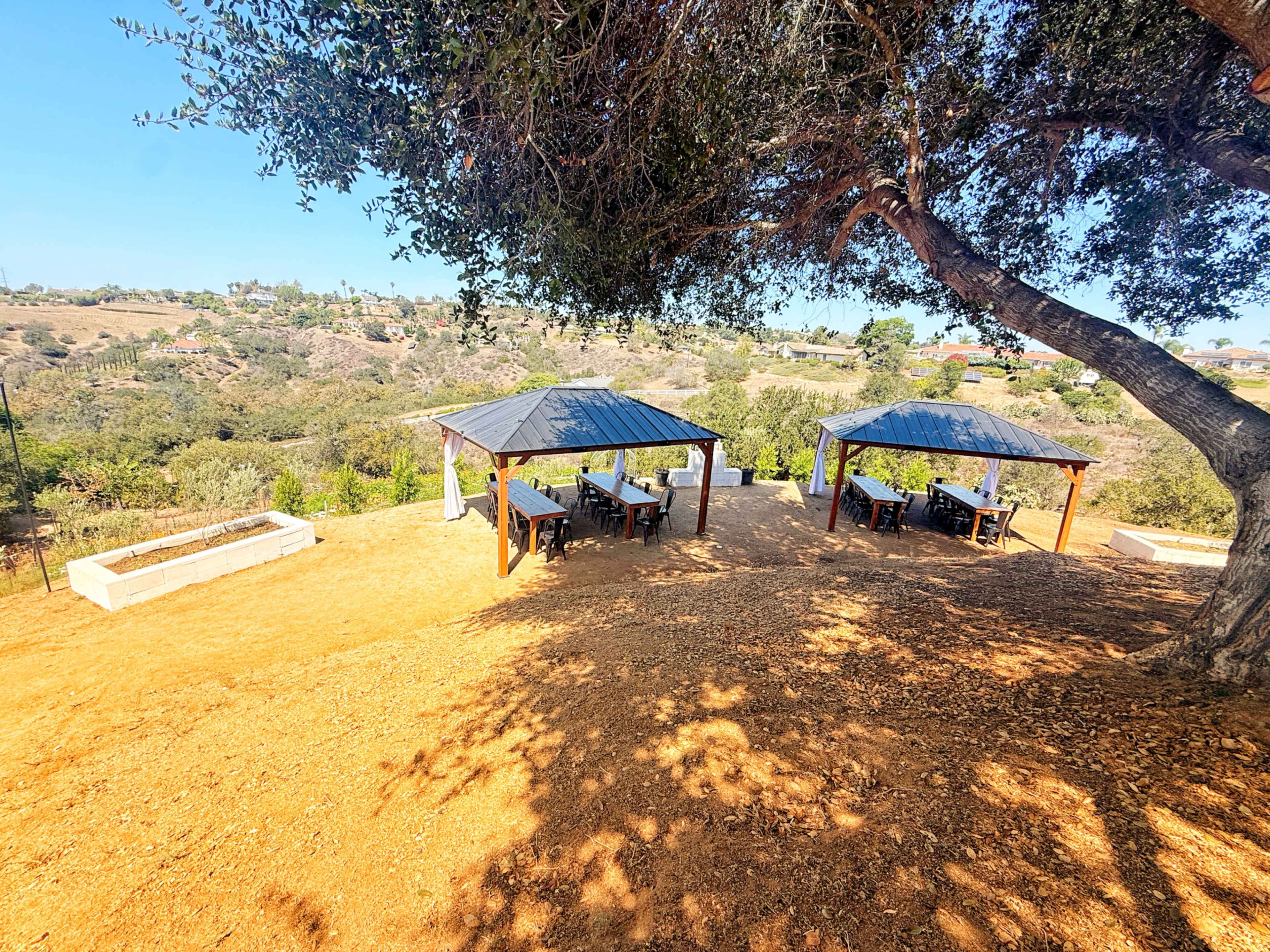 The image shows two wooden gazebos with tables and chairs, situated on a hillside overlooking a green valley.