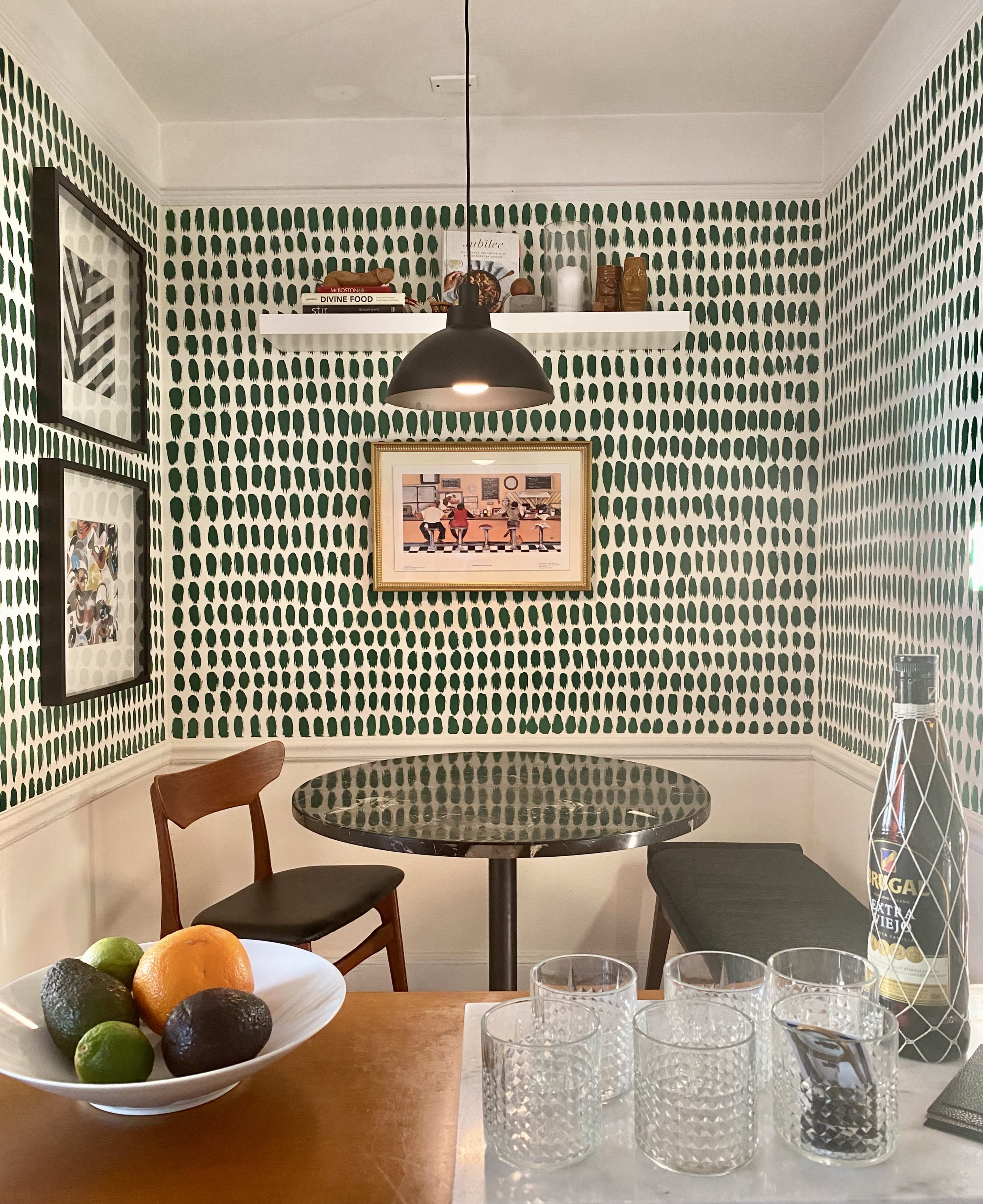A small dining nook features a round glass table with two chairs, surrounded by green dotted wallpaper and adorned with a shelf displaying a framed picture.