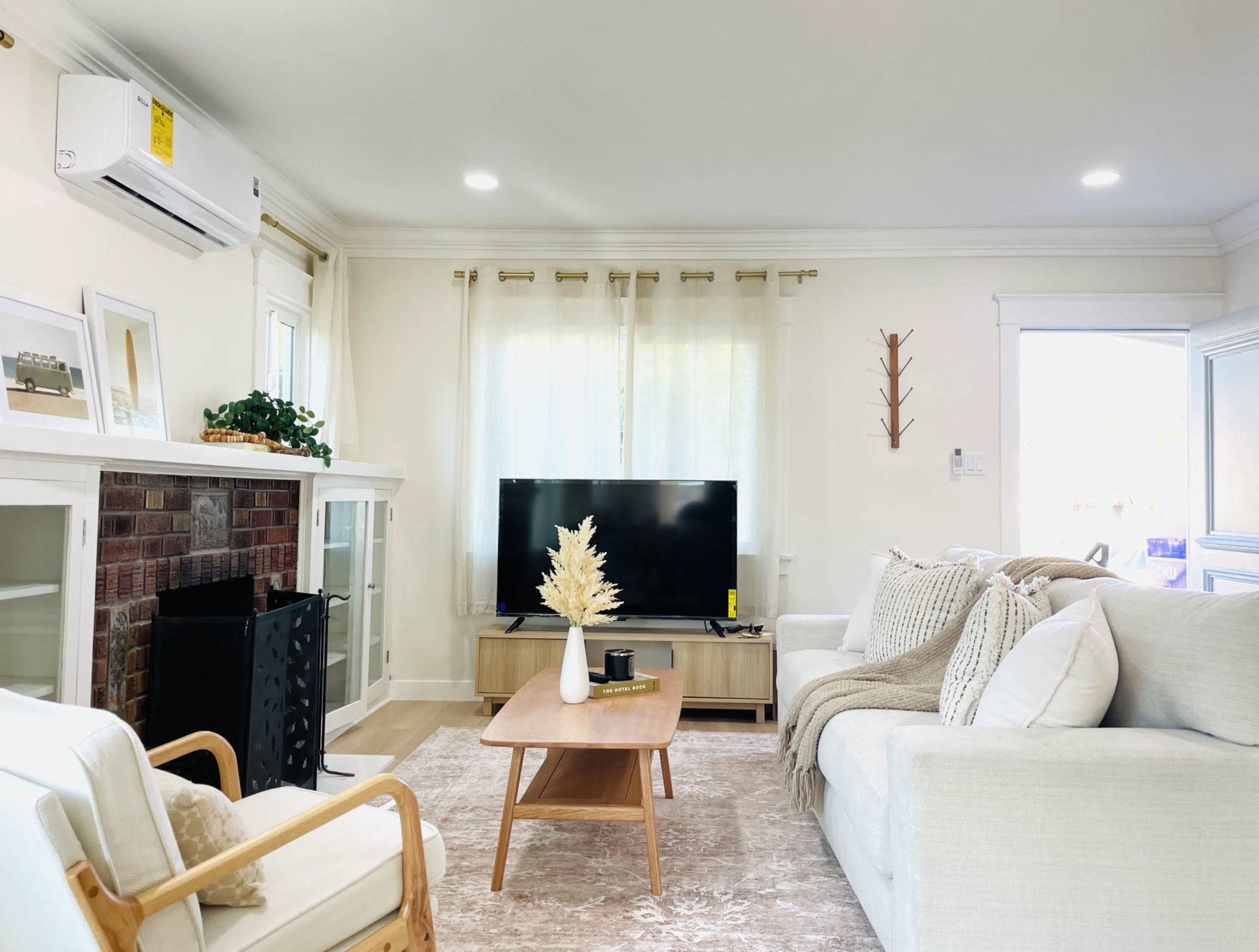 A bright living room with a white sofa, a wooden coffee table, a television on a stand, and a fireplace adorned with decorative elements.