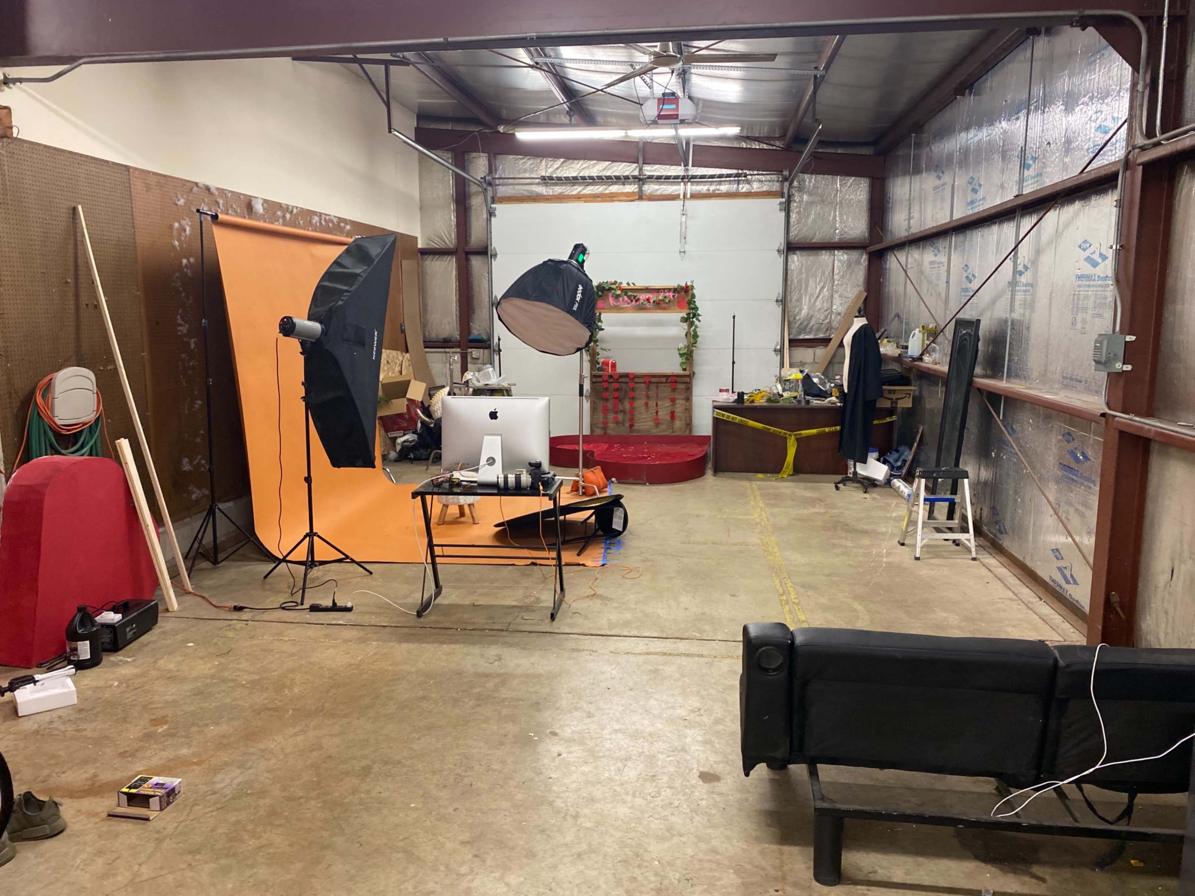 A spacious room with photography equipment set up, including an orange backdrop, softbox lights, and a computer on a table, surrounded by various props and furniture.
