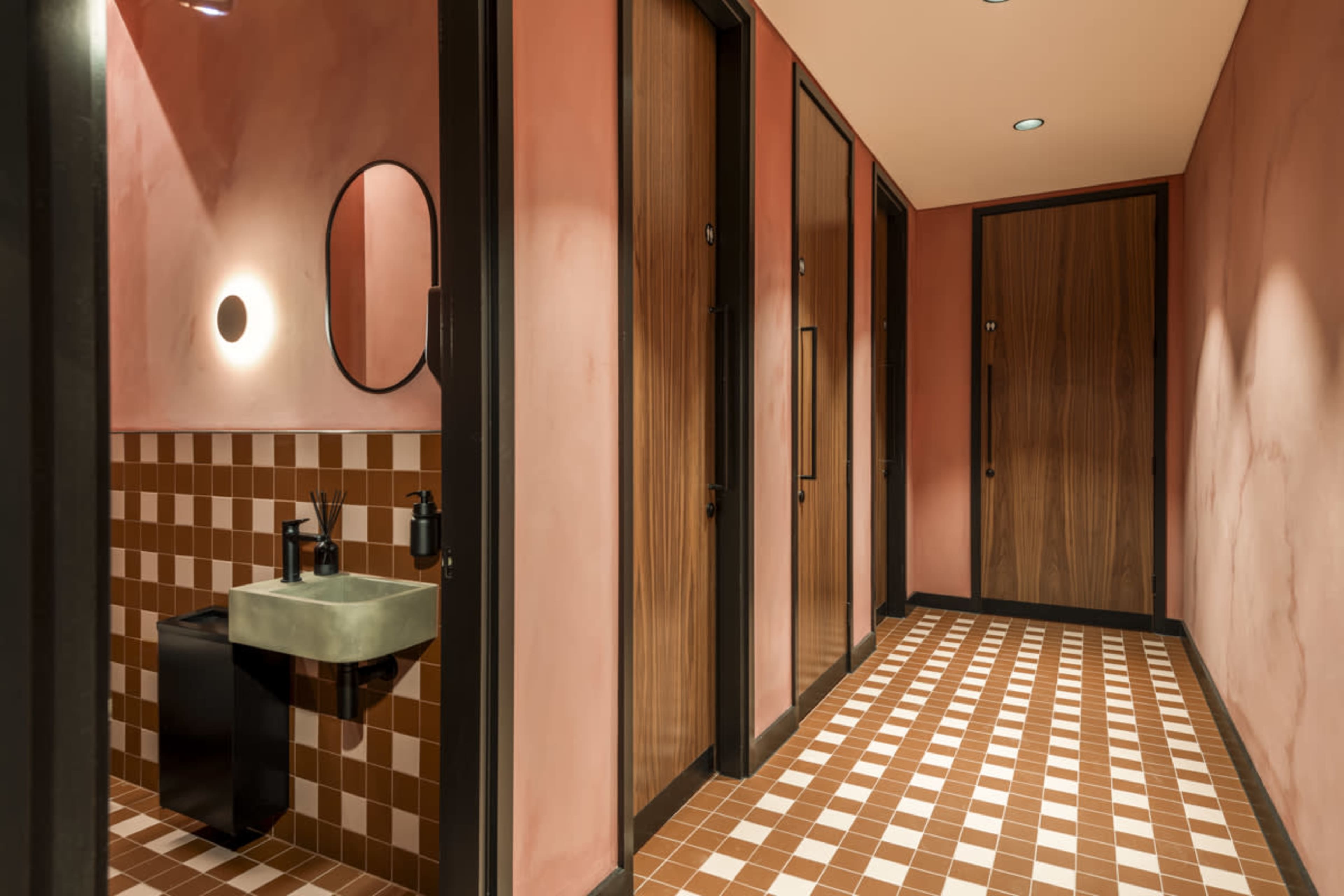 The image depicts a modern restroom with pink walls, a tiled floor, and several closed wooden doors lining the hallway.