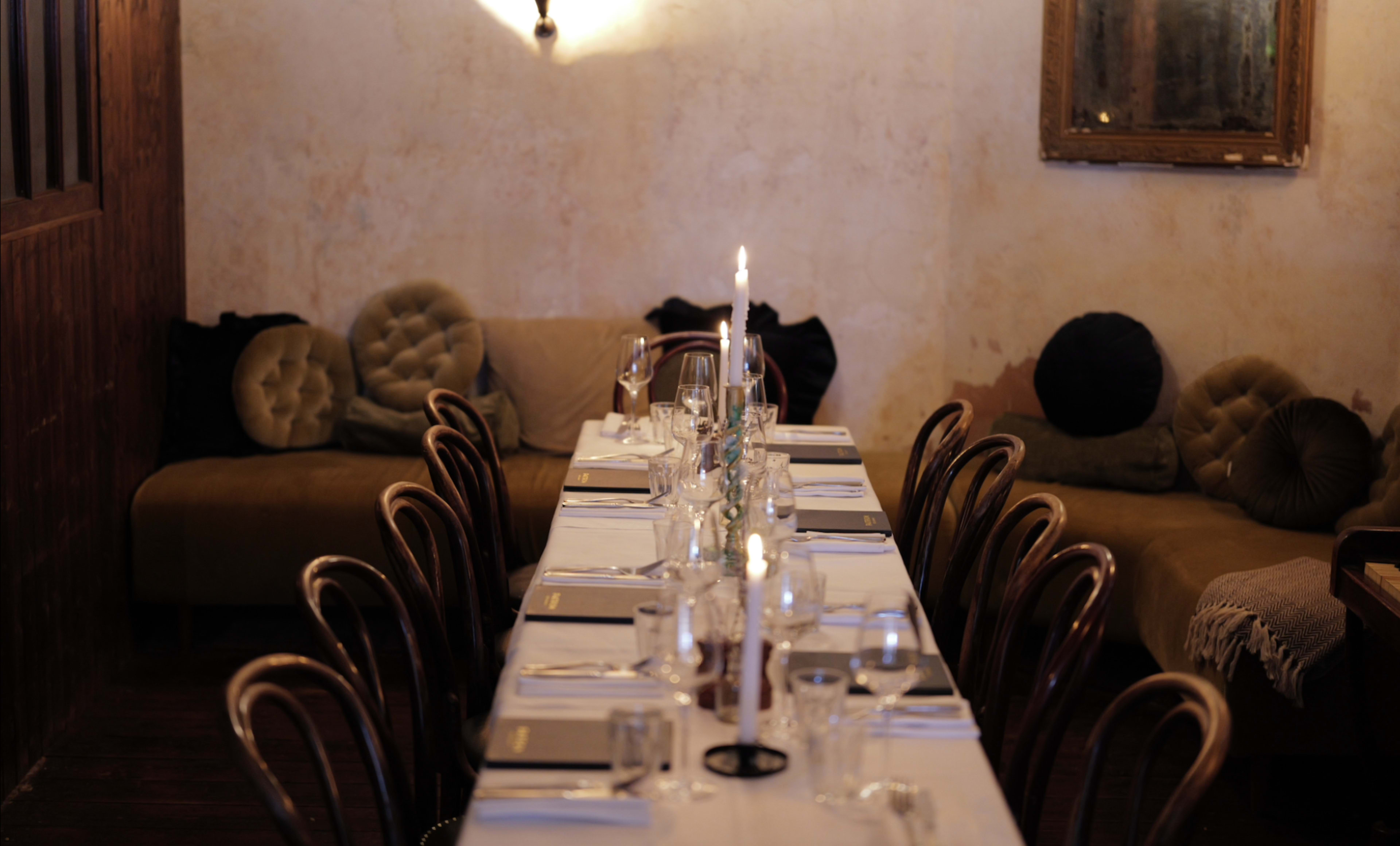 A long dining table is set with glassware and cutlery, illuminated by candlelight, surrounded by wooden chairs in a softly lit room.