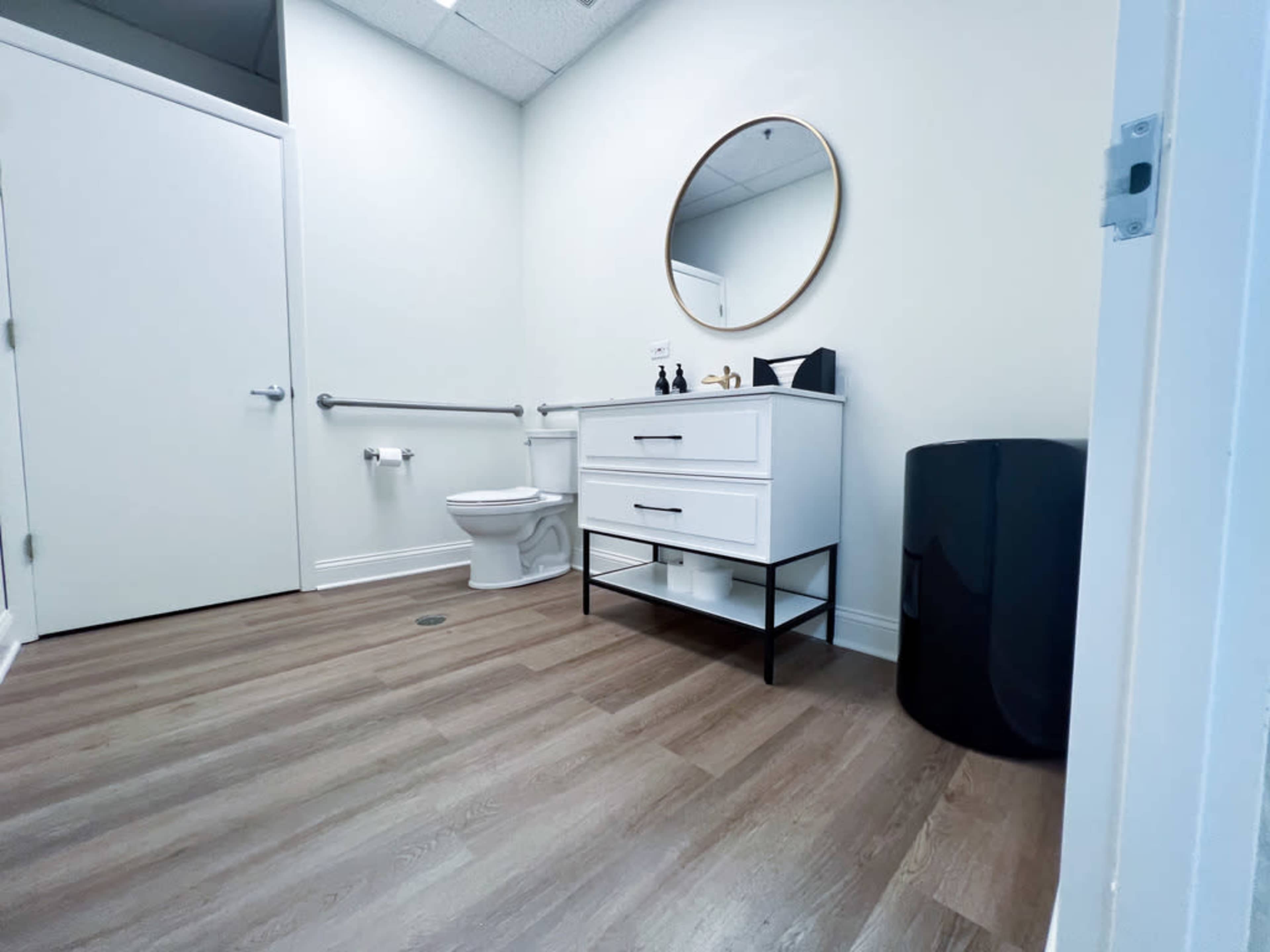 A clean restroom features a white vanity with a round mirror, a toilet, and a black wastebasket against light-colored walls and wooden flooring.