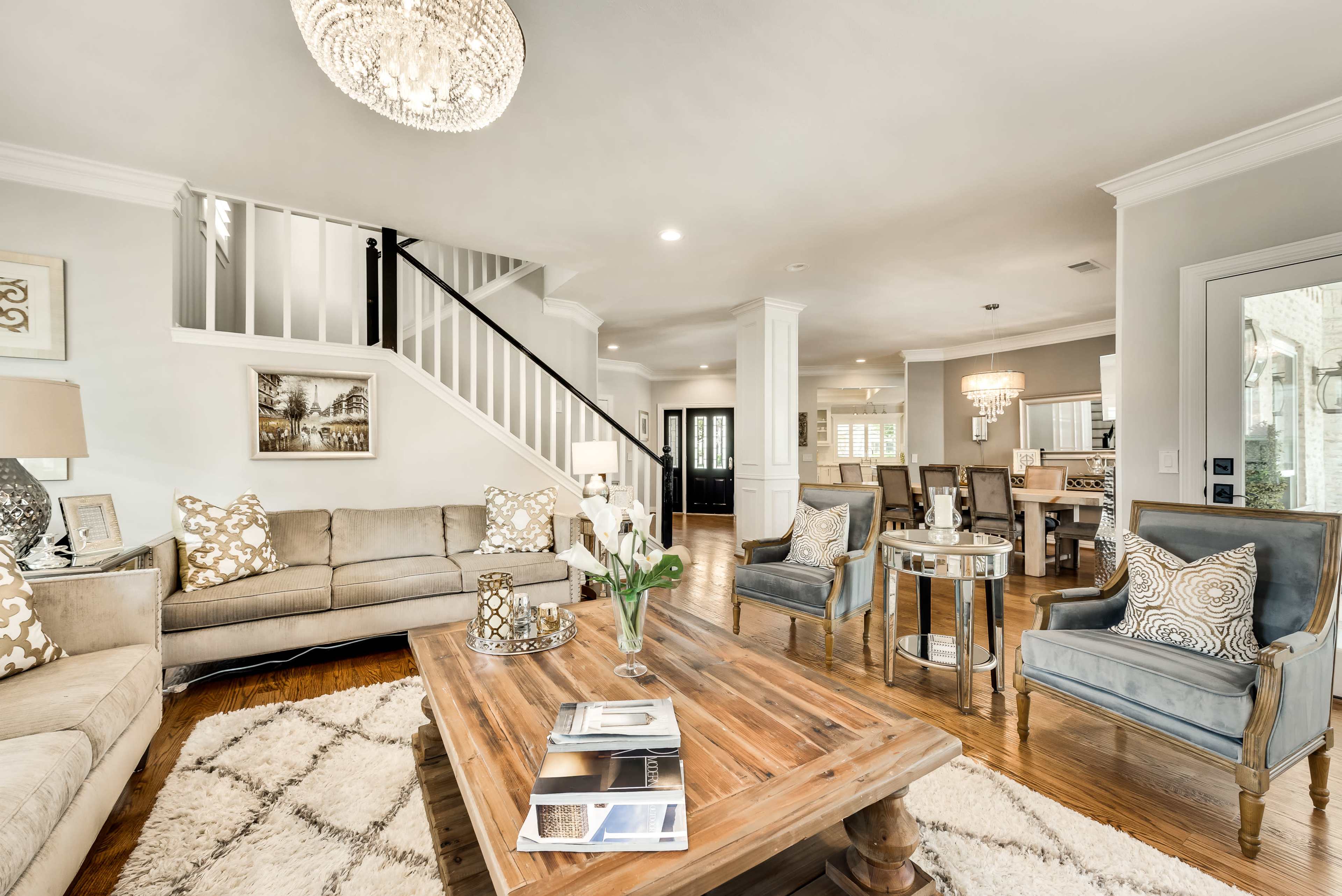A spacious living room features a central wooden coffee table surrounded by a sofa, two patterned armchairs, and a decorative chandelier above, with a staircase visible in the background.