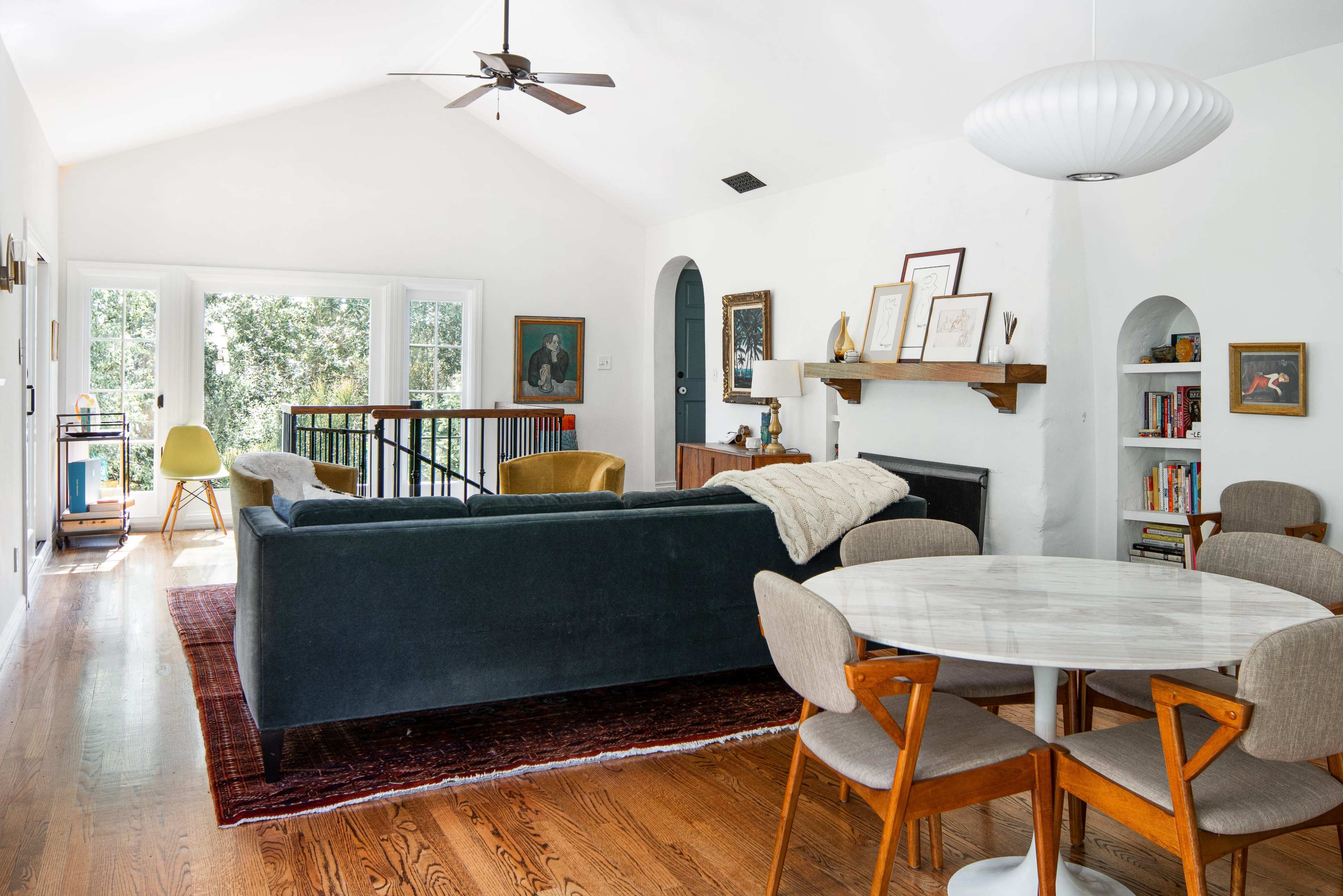 A bright, open living space features a dark green sofa, a round marble dining table with wooden chairs, and large windows overlooking a landscaped area.