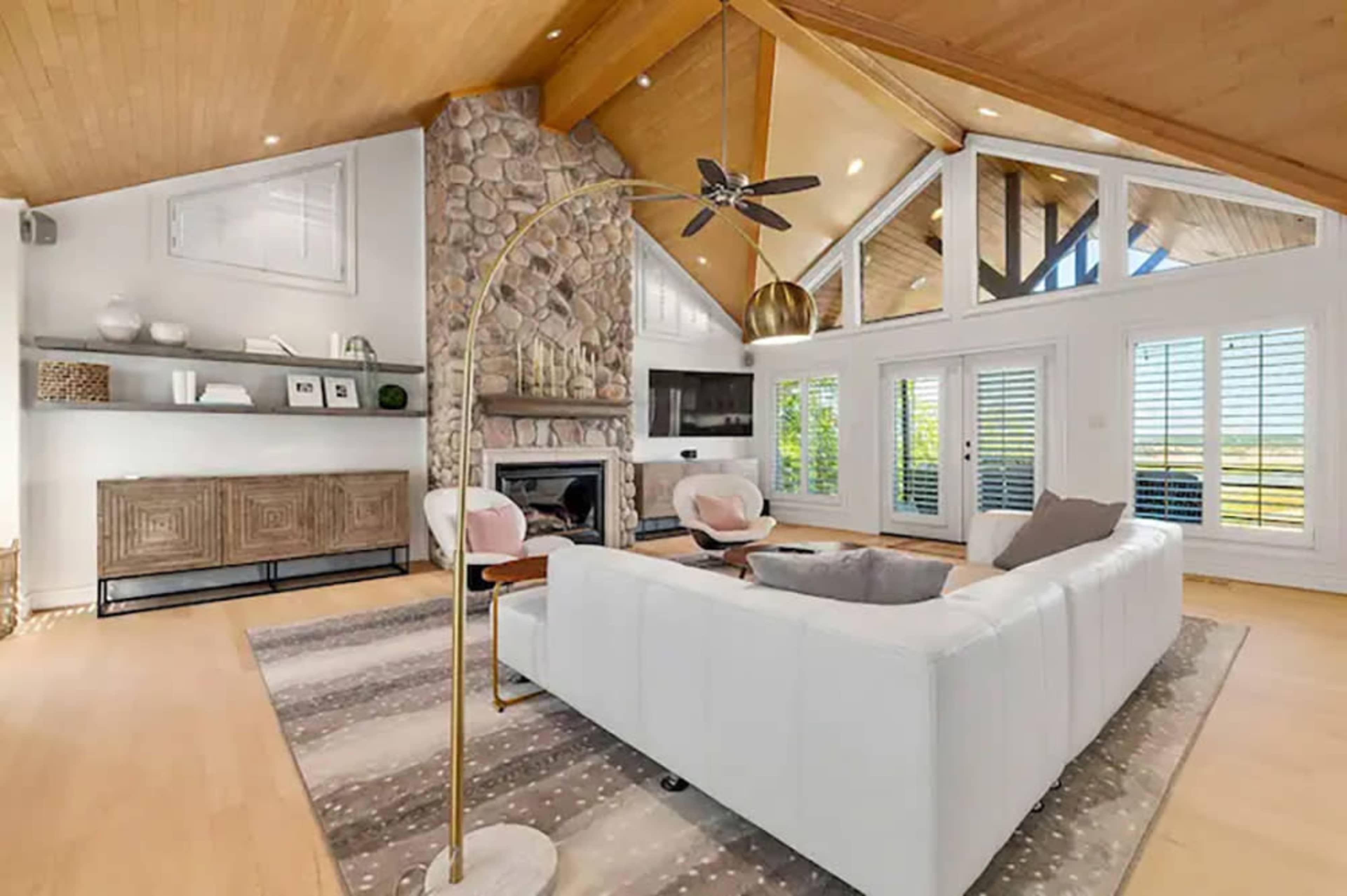 The image shows a modern living room with a stone fireplace, large windows, and white furniture arranged around a rug.