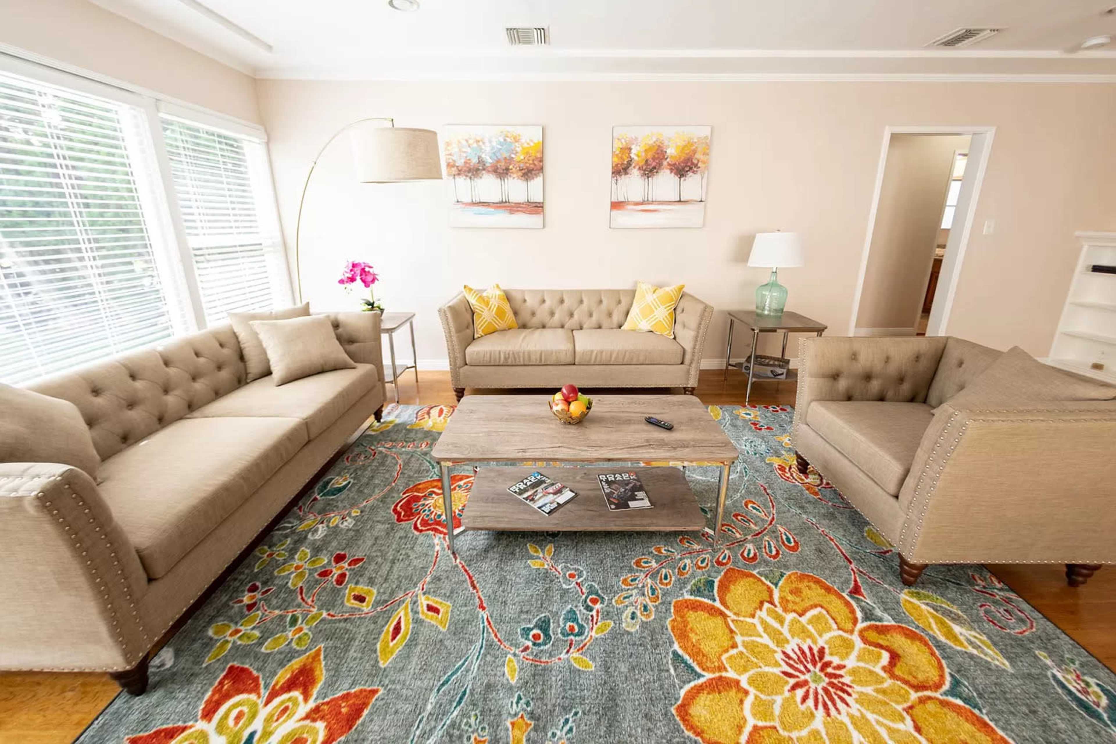 The image shows a cozy living room furnished with two beige tufted sofas, a wooden coffee table, and a colorful patterned rug, with natural light streaming through large windows.