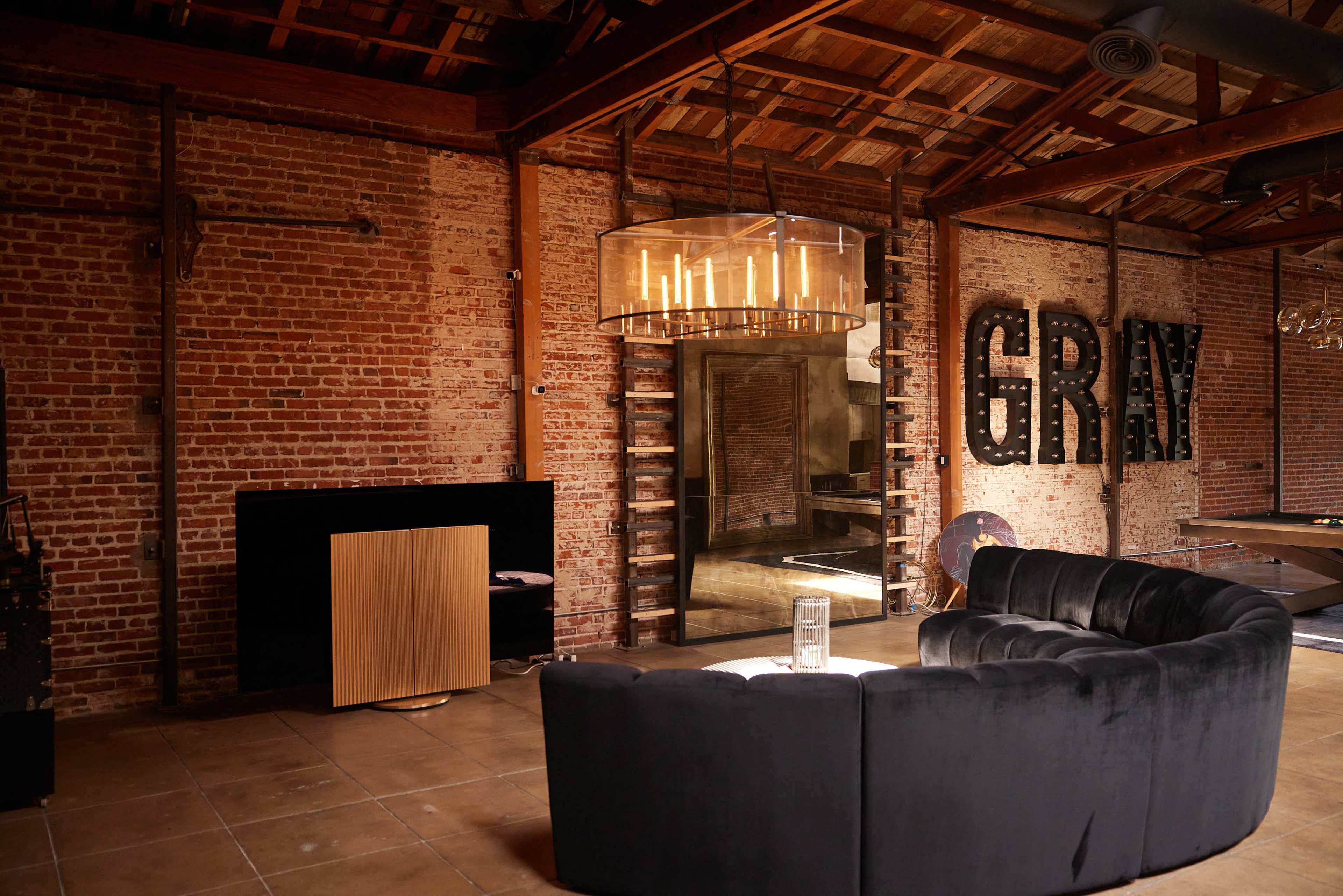 The image shows a modern lounge area featuring a curved, dark sofa, a large chandelier, and exposed brick walls with the word "GRAY" displayed prominently.