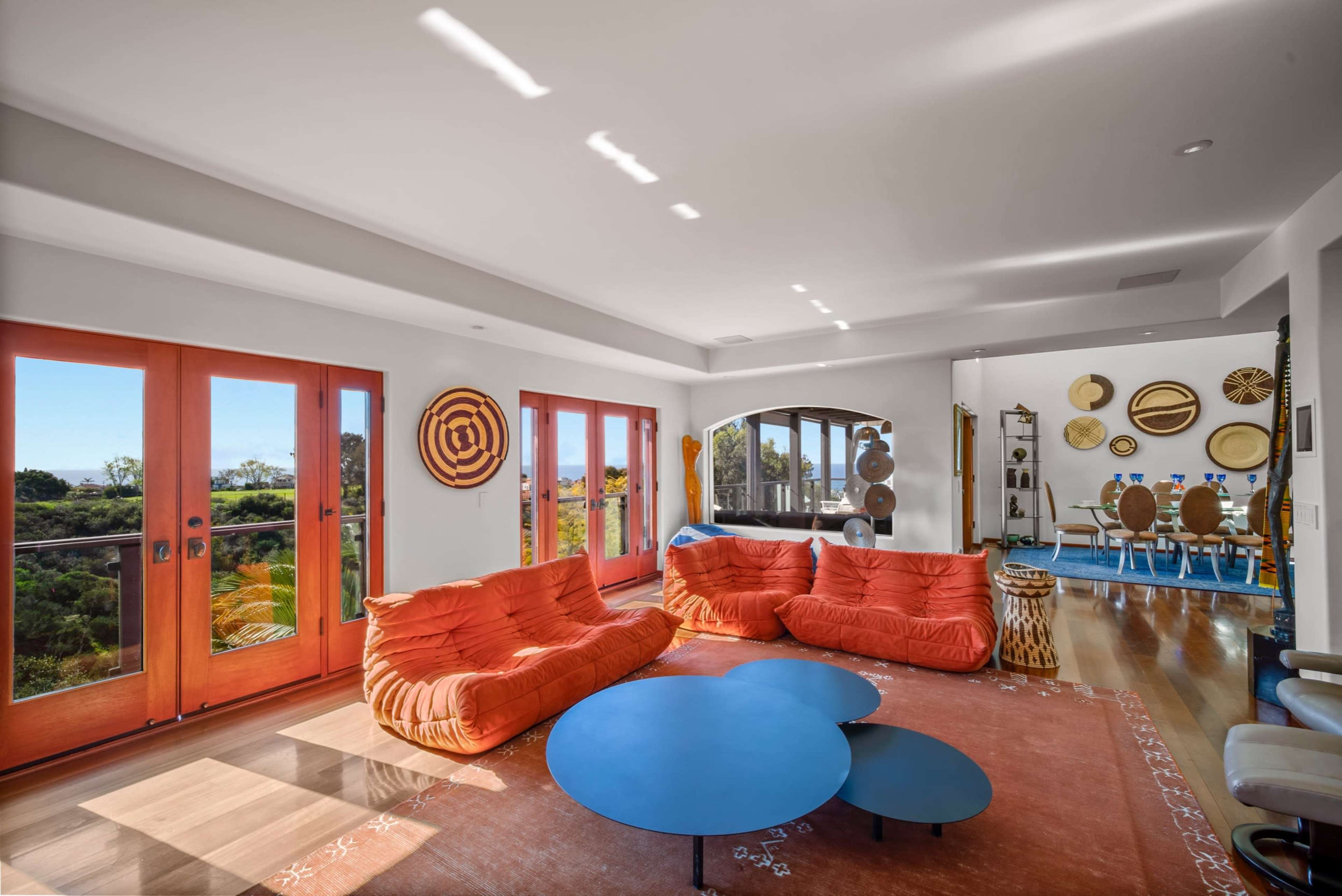 A bright living room features orange sofas, a round blue coffee table, and large windows opening to a scenic view.