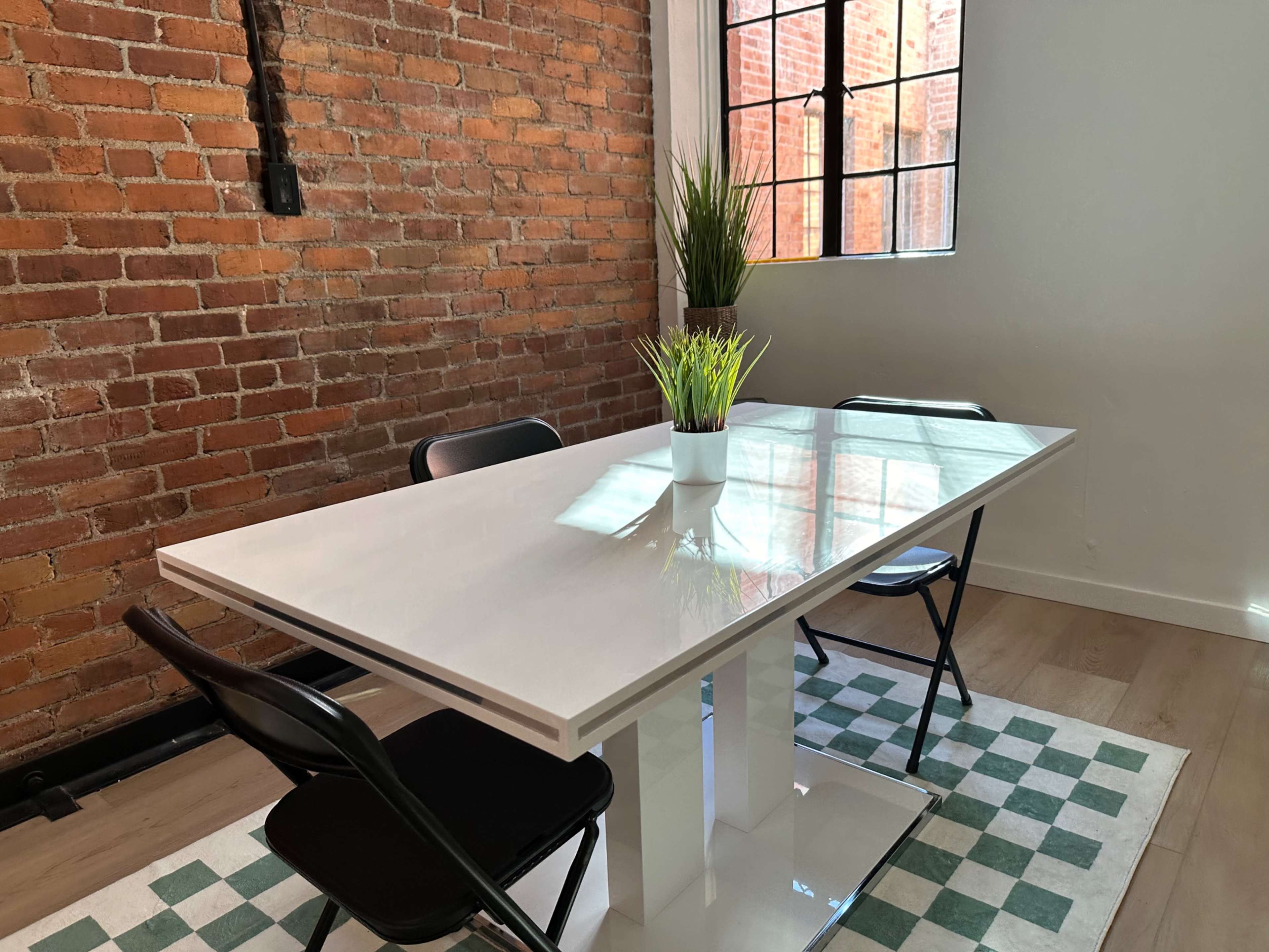 A white dining table with two black chairs sits on a checkered green and white floor, illuminated by natural light from a window against a brick wall.