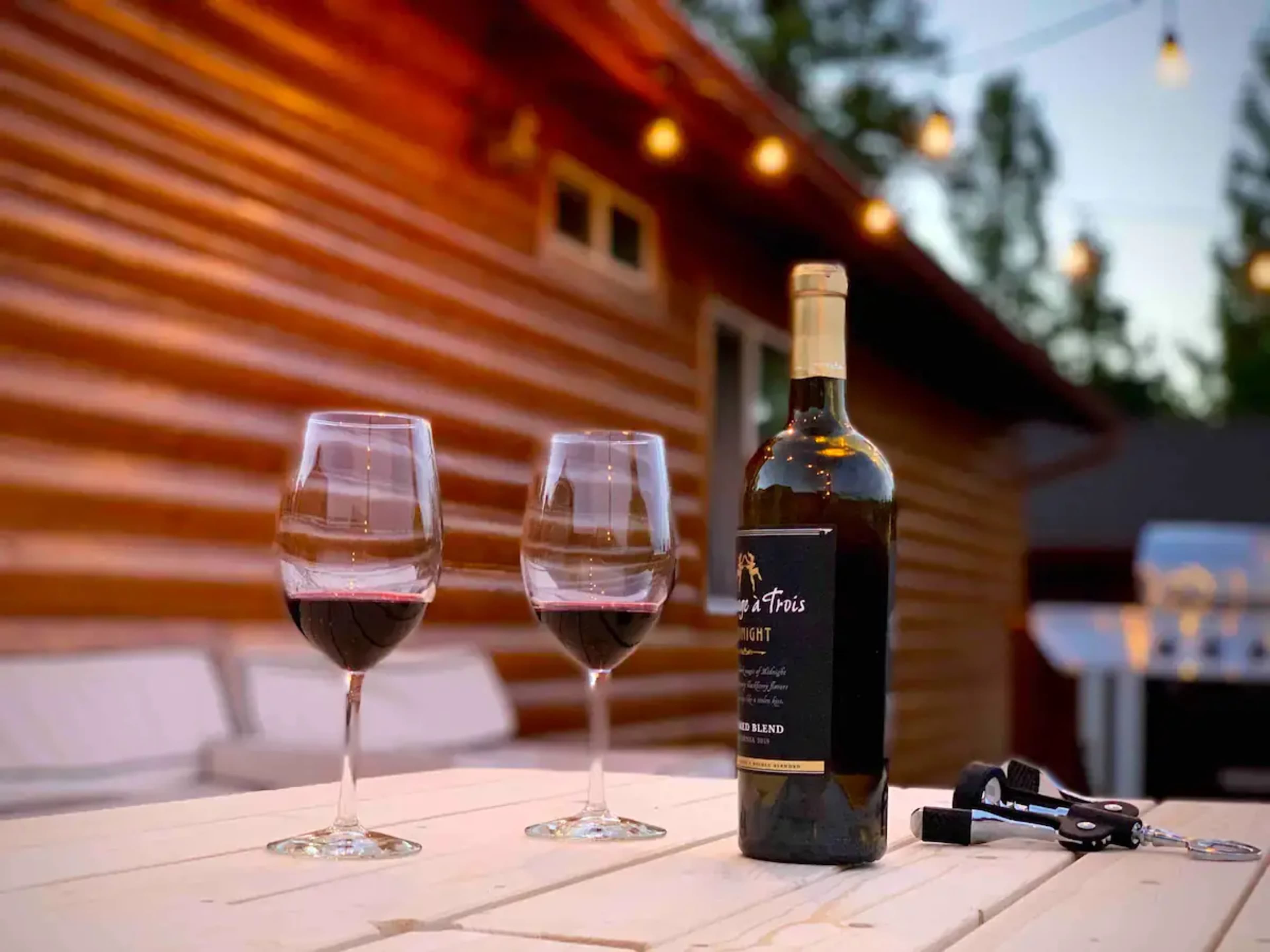 Two glasses of red wine sit next to an open bottle on a wooden table, with string lights illuminating a rustic cabin background.