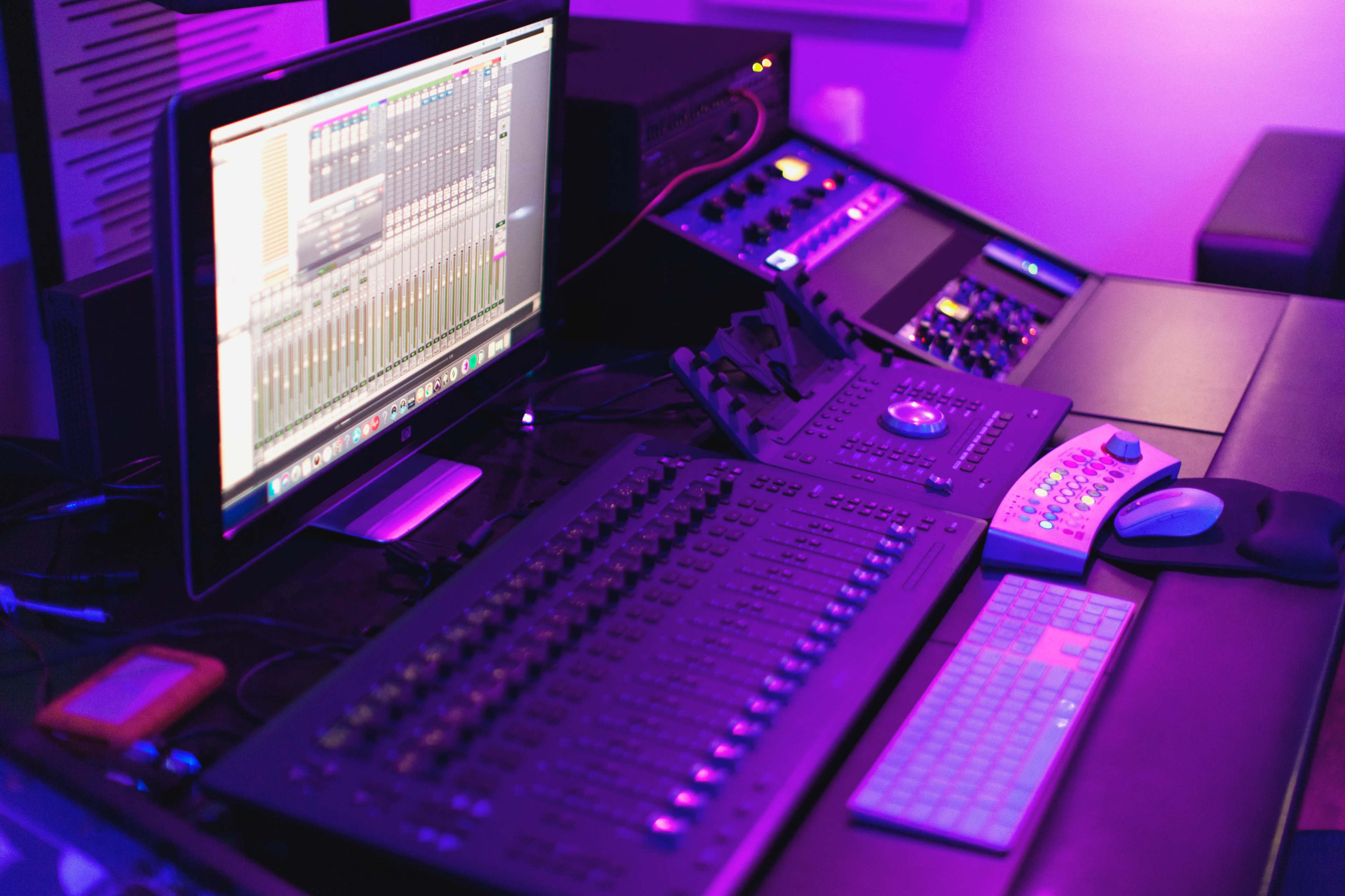 A music production workspace featuring a computer monitor displaying audio mixing software, a control surface with faders, and various sound equipment in a dimly lit setting with purple lighting.