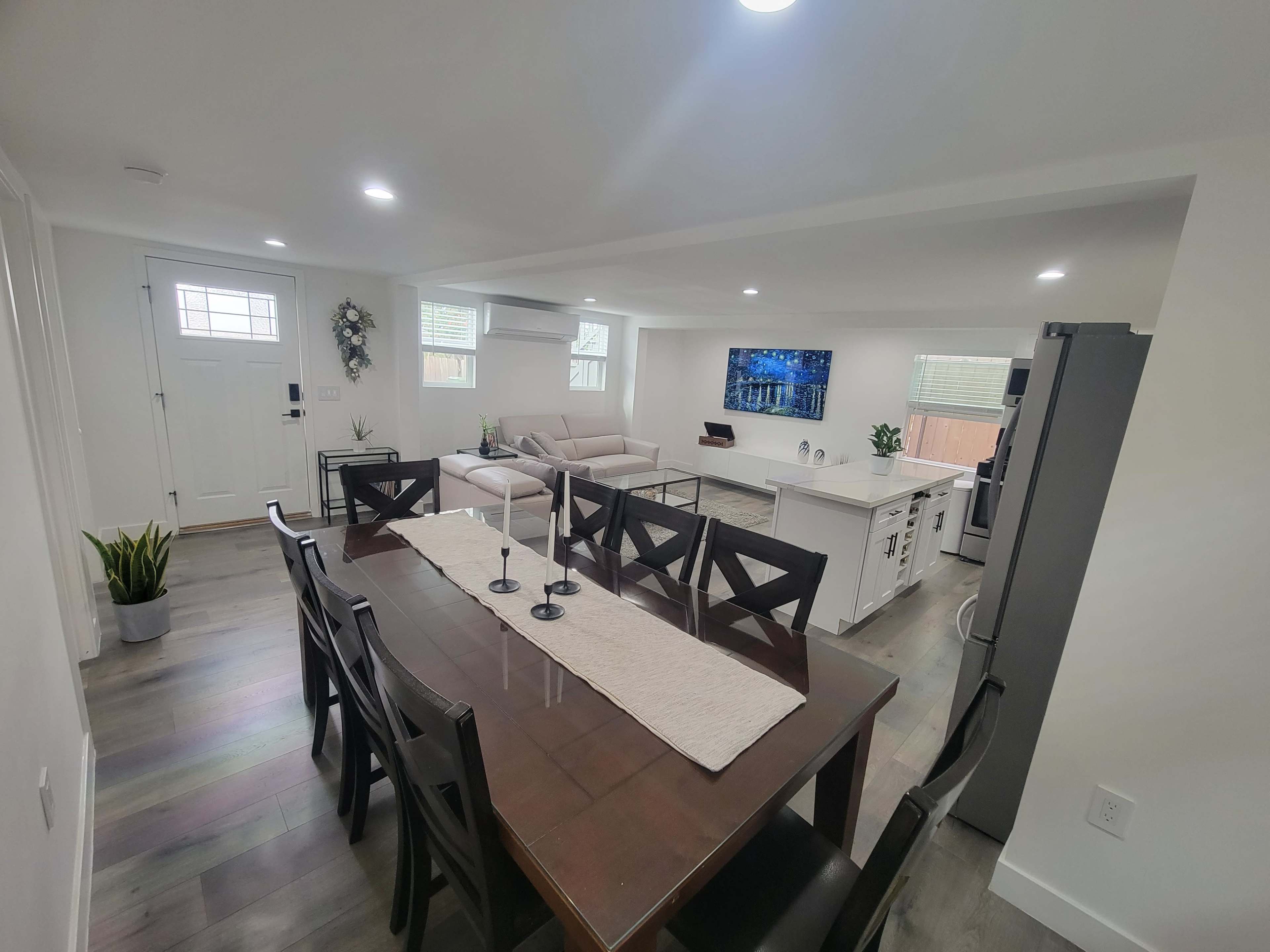 A modern open-concept living and dining area features a dining table, a sofa, a wall-mounted television, and a kitchen island.