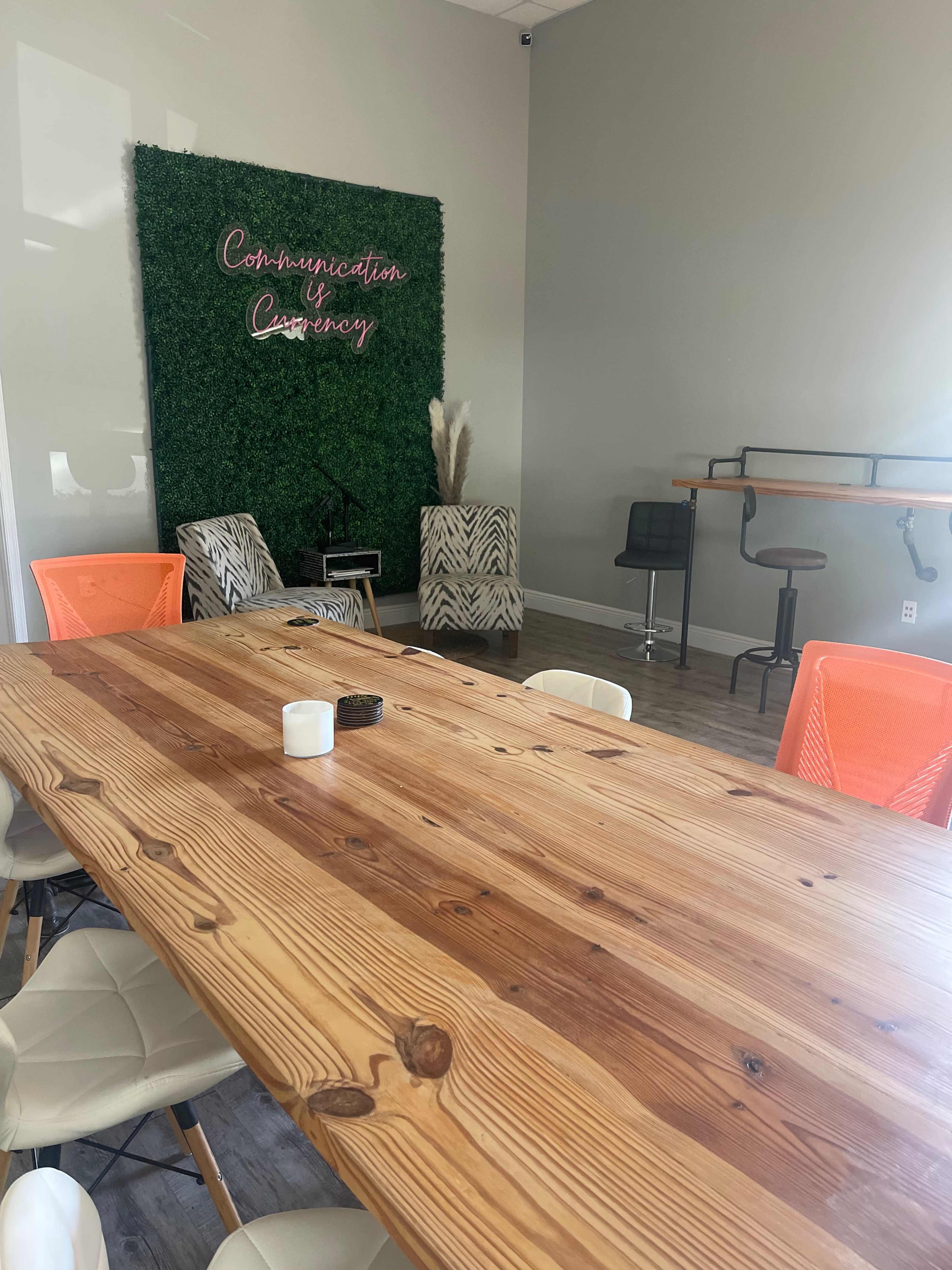 A modern meeting space featuring a large wooden table surrounded by white and orange chairs, with green wall decor displaying the phrase "Communication y Currency."