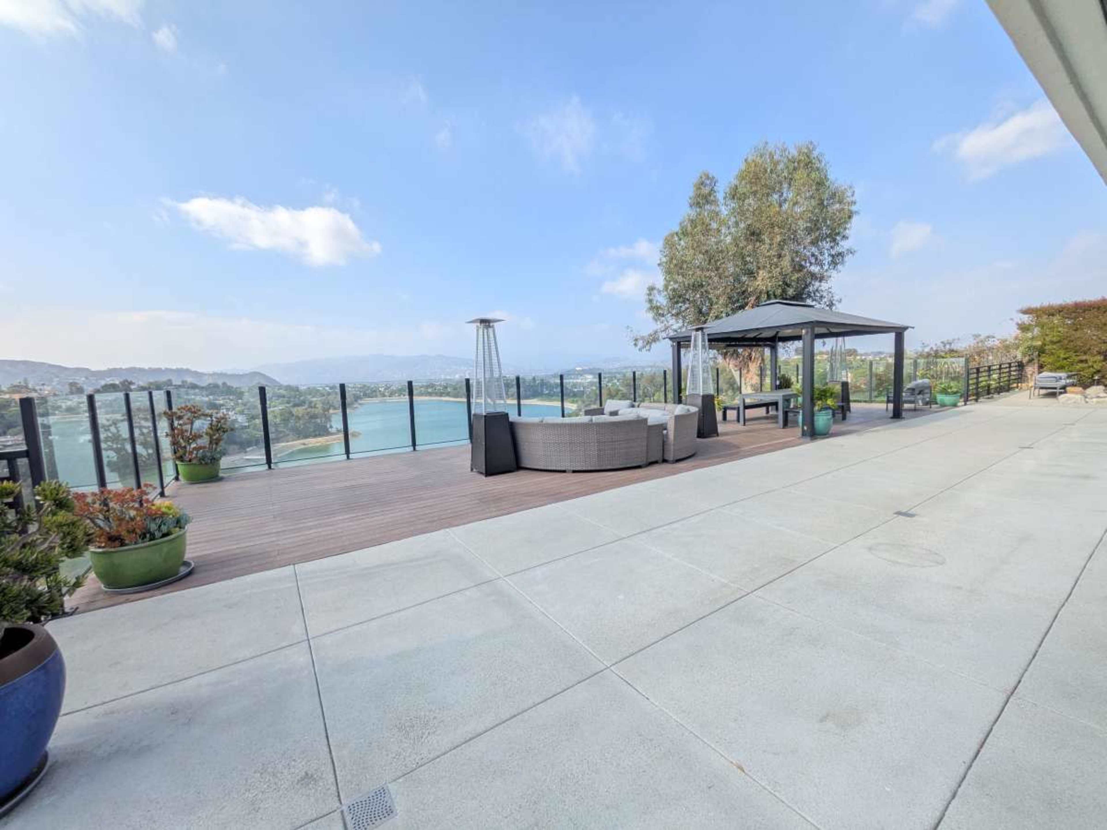 The image shows a spacious outdoor patio with a seating area, a gazebo, and a view of a lake surrounded by hills under a clear sky.