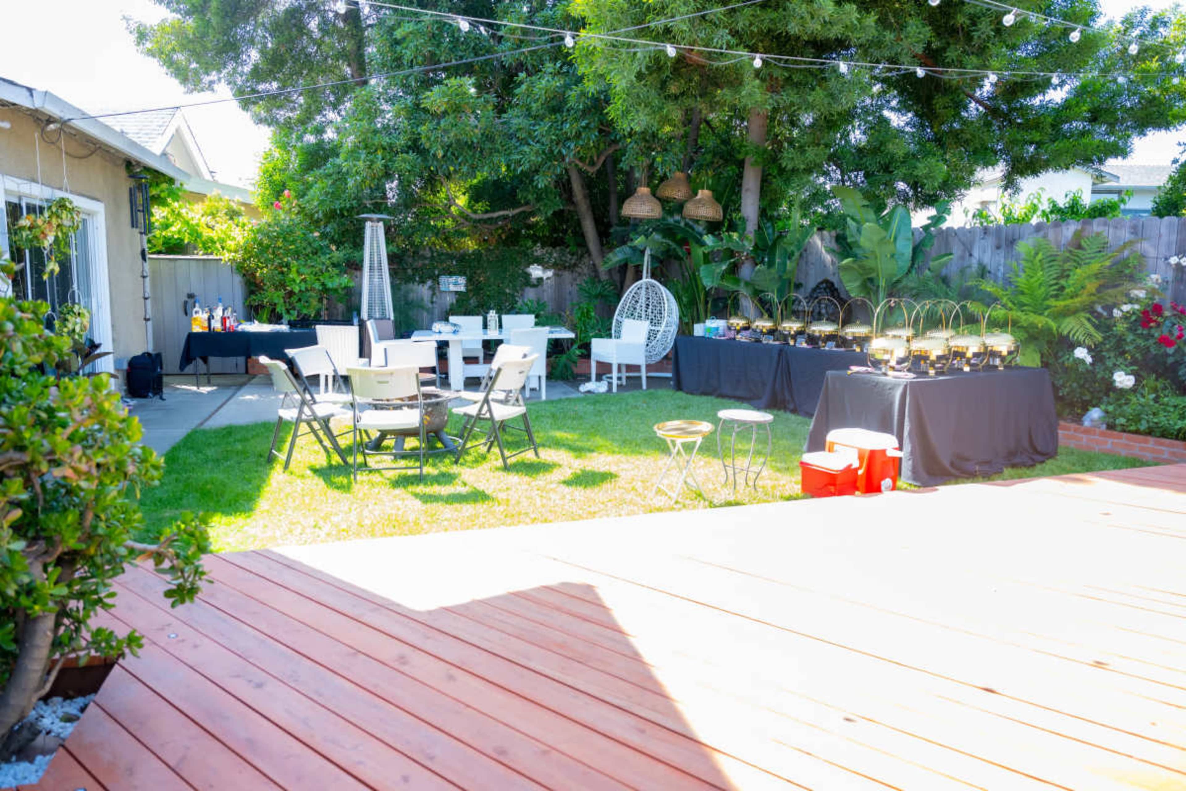A backyard party setup features tables with black cloths, chairs, and decorative elements surrounded by greenery and string lights.