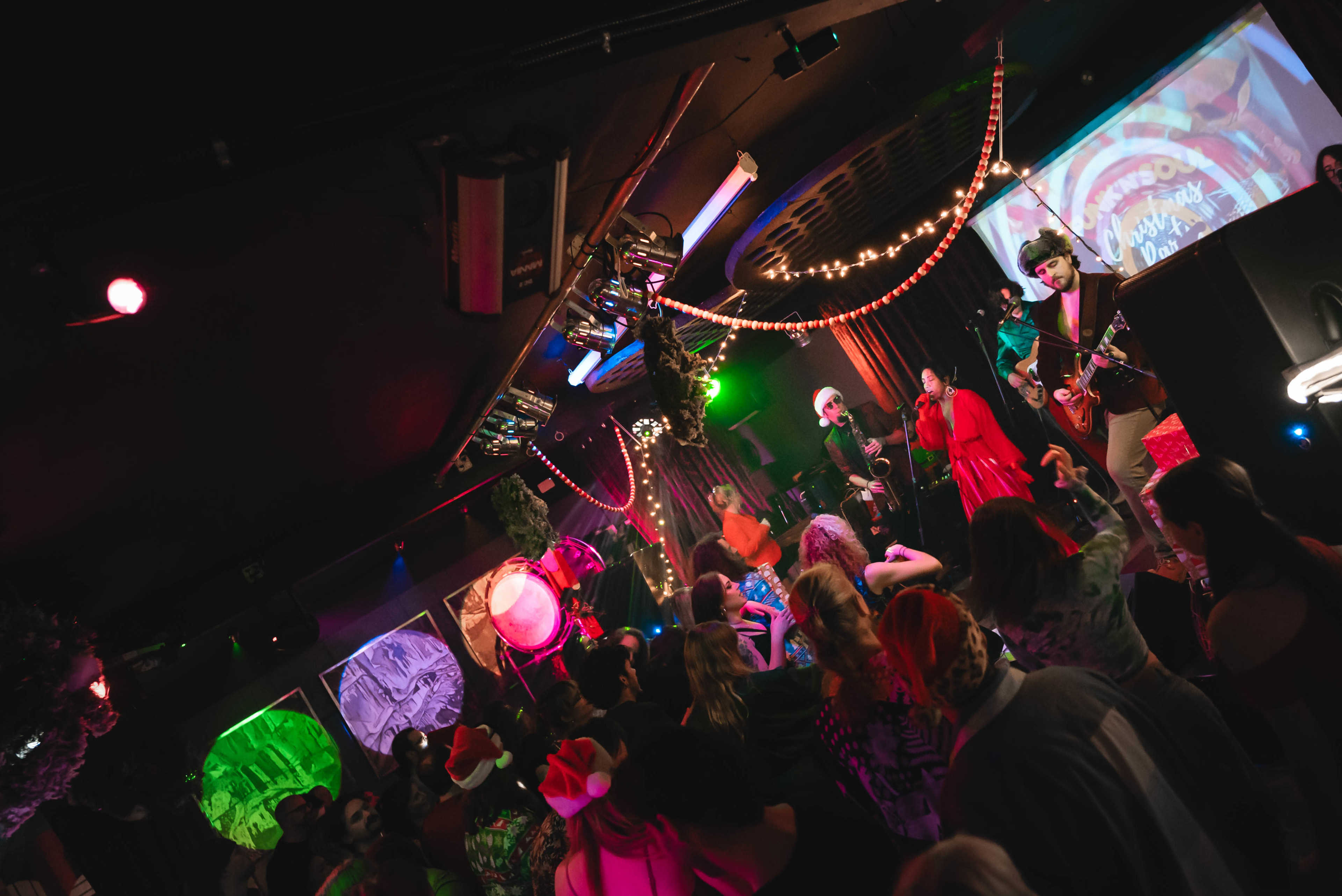 A lively crowd dances in a dimly lit venue decorated with colorful lights and visual projections during a performance.