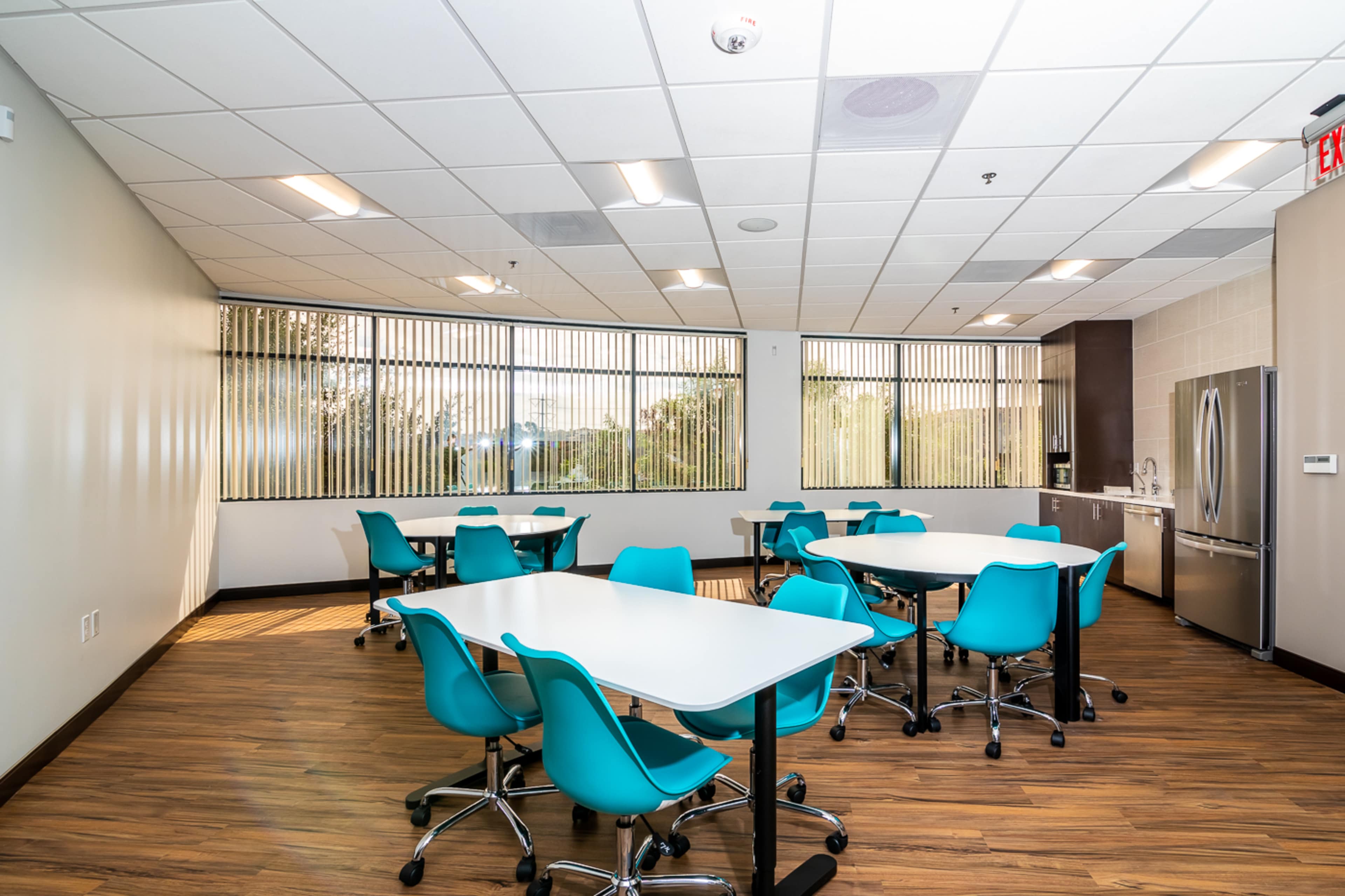 The image shows a modern dining area with multiple white tables and turquoise chairs, lit by natural light through large windows.