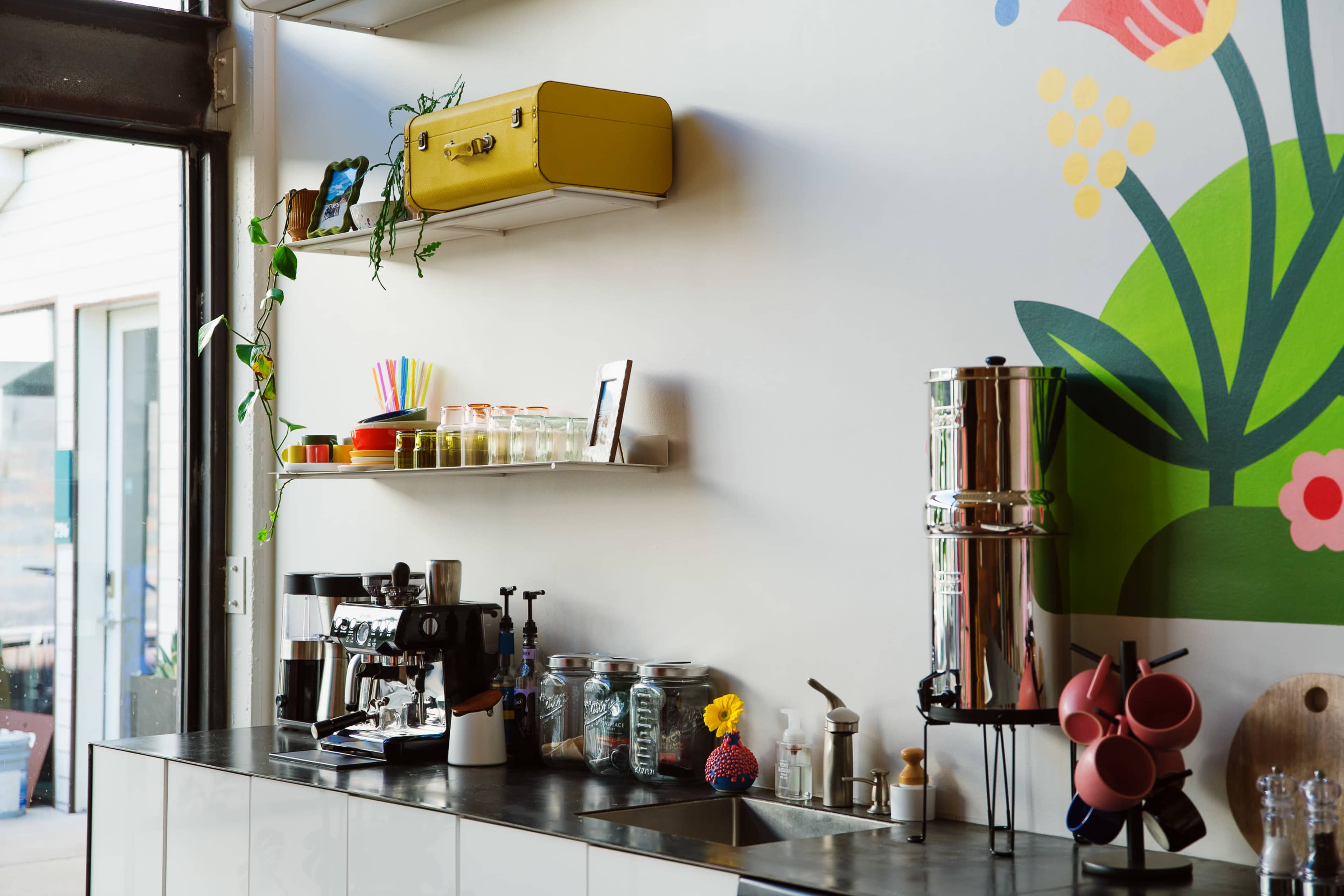 The image shows a modern kitchen area featuring a coffee machine, various glass jars, and a yellow suitcase shelf against a colorful mural with flowers.