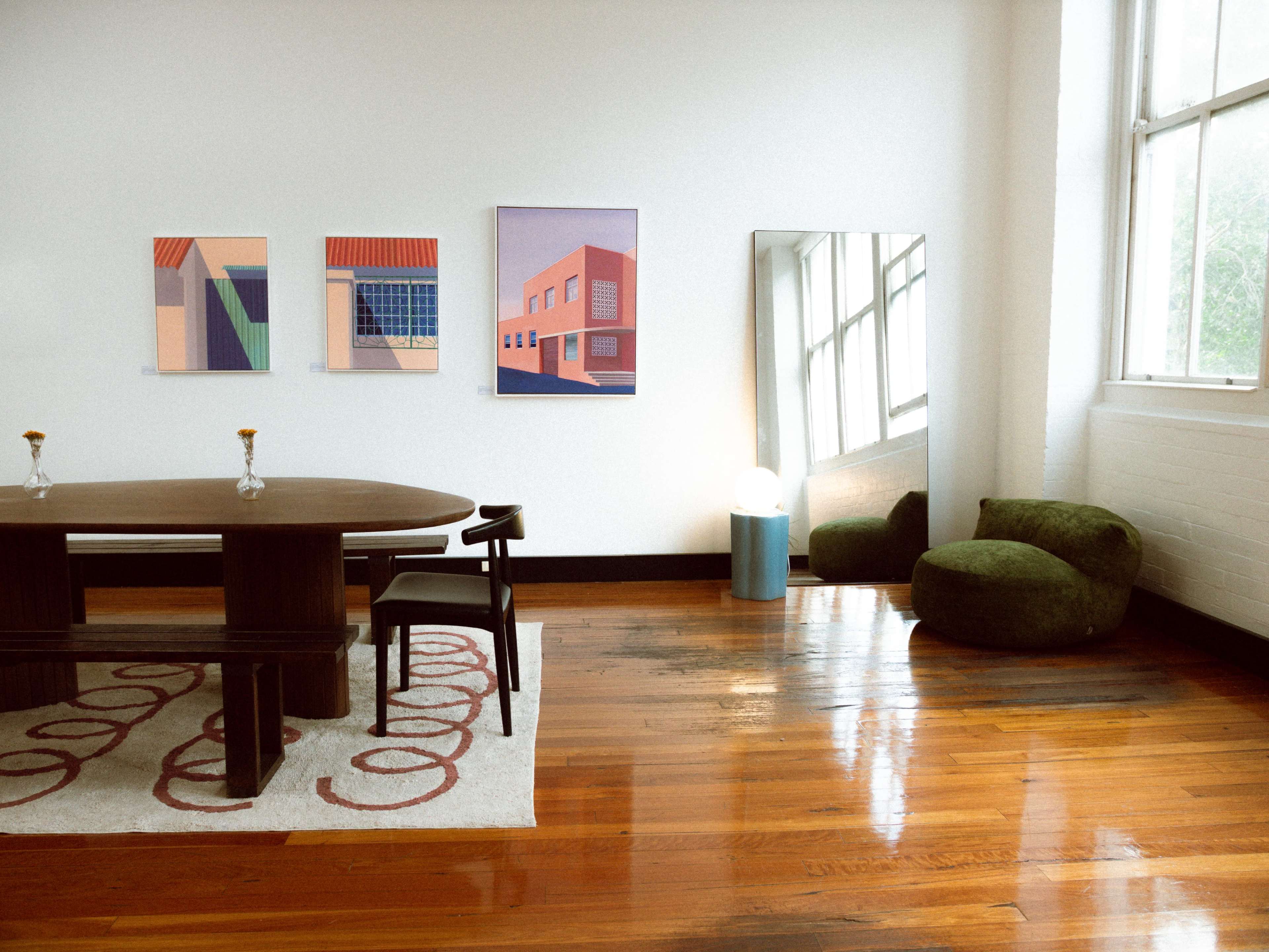 The image shows a modern interior room featuring a dining table, four framed artworks on the wall, a large mirror, a round green chair, and polished wooden floors.