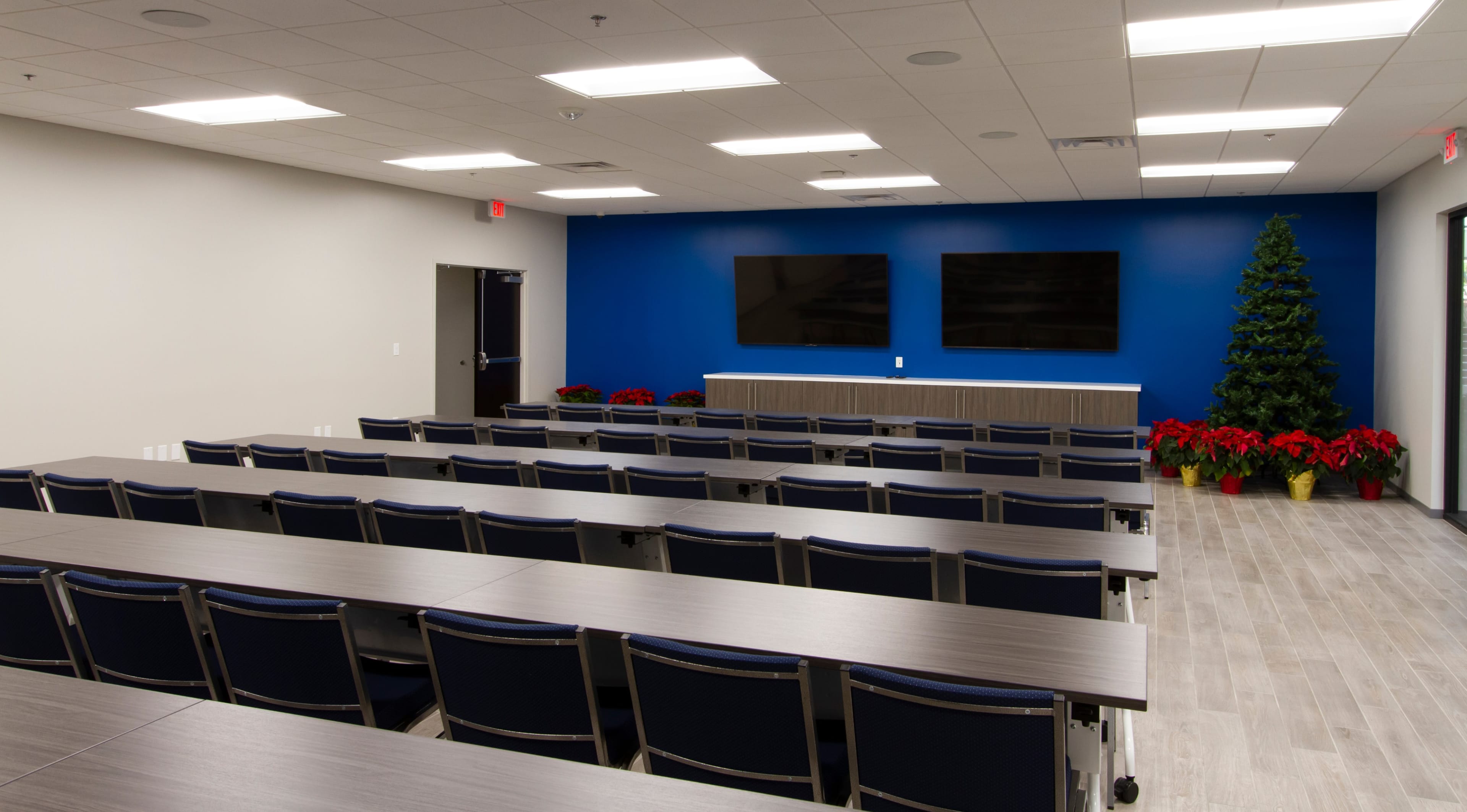 A conference room with rows of tables and chairs, two large screens on a blue wall, and a decorative tree in the corner.