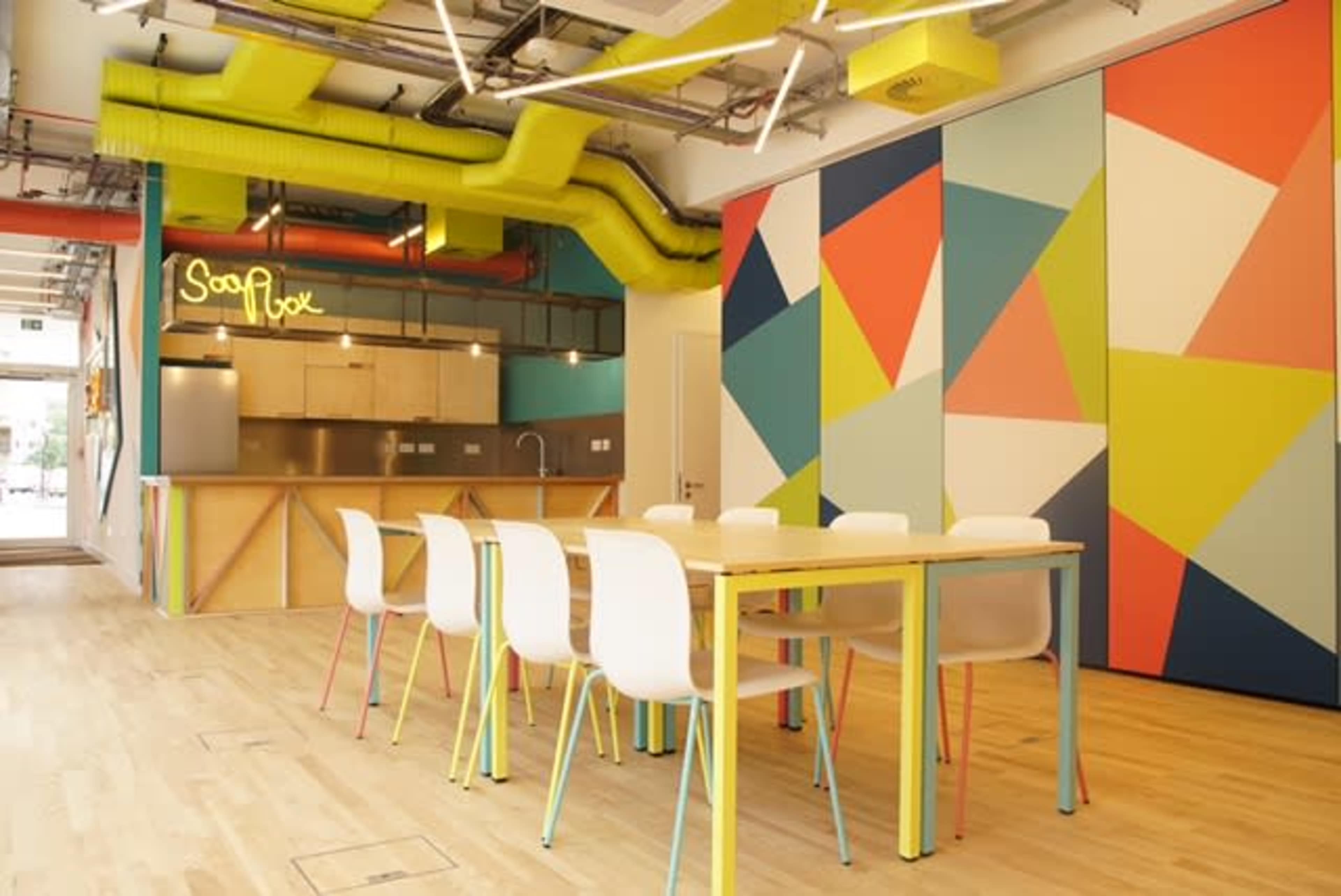 A modern kitchen and dining area featuring a long table with white chairs, colorful geometric wall designs, and a bright green ventilation system.