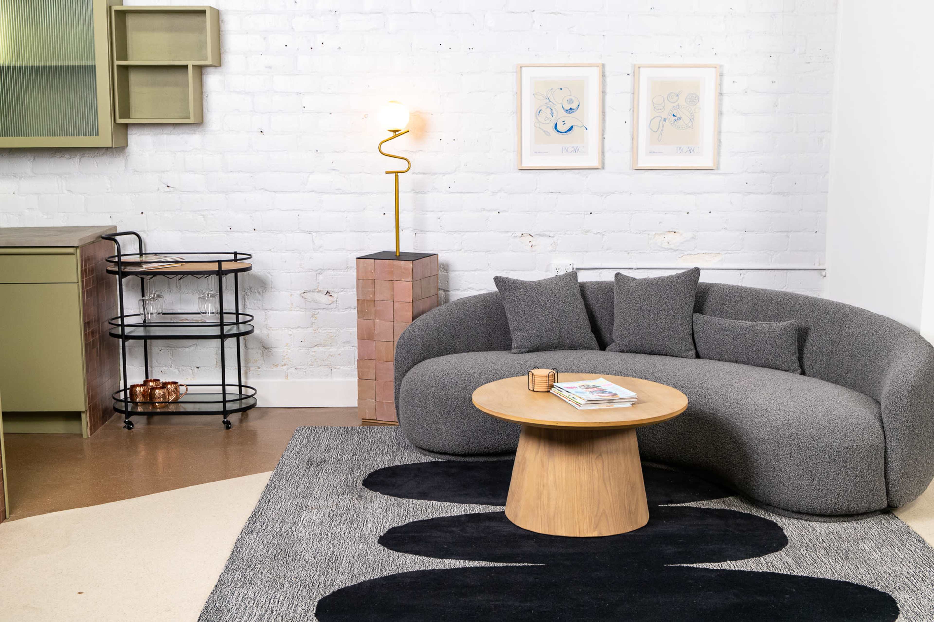 A modern living room features a curved gray sofa, a circular wooden coffee table, and a patterned black rug, with minimalist art on the brick wall and a standing lamp beside a cabinet.