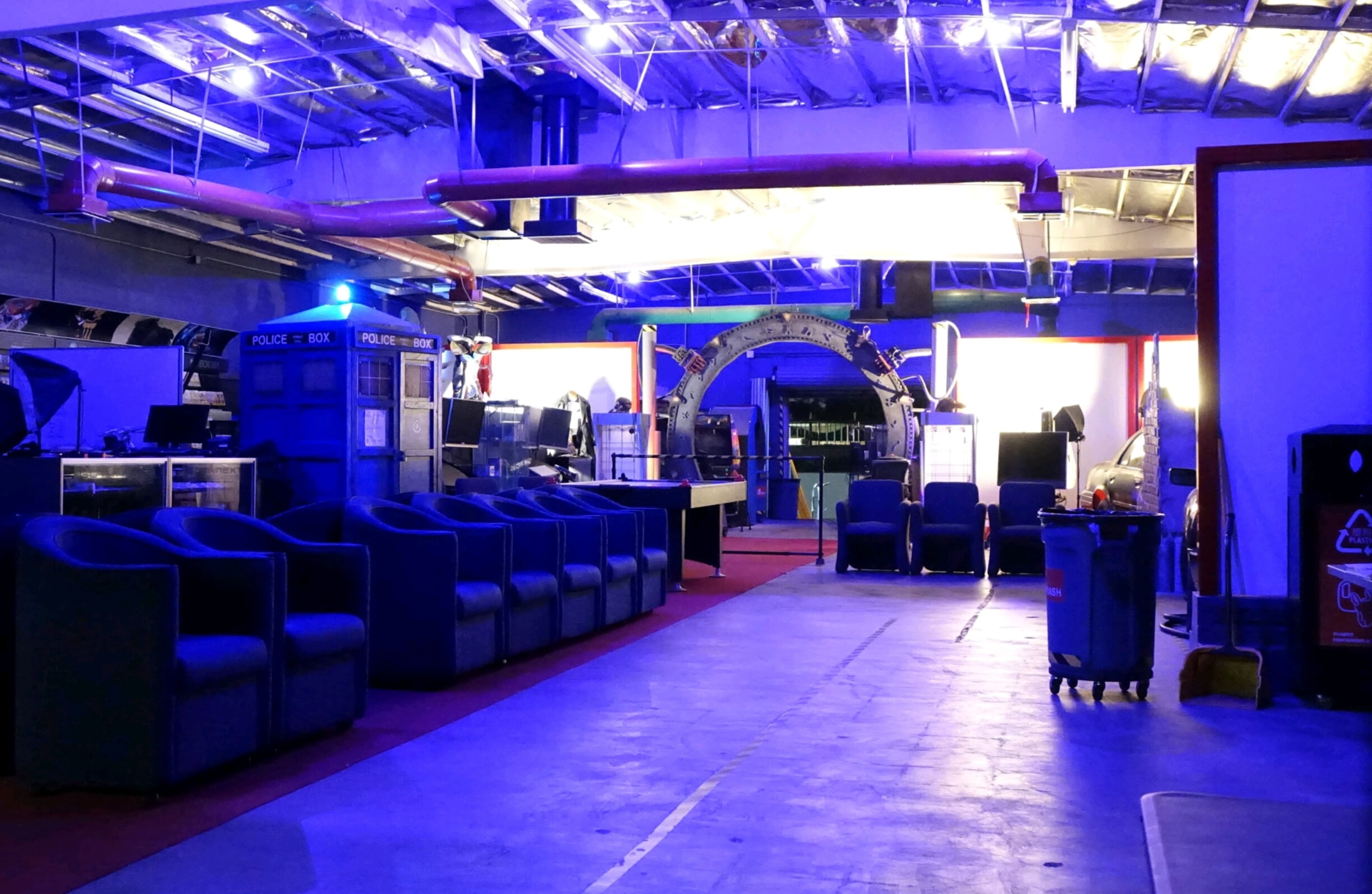 A dimly lit room features blue lighting, an arrangement of chairs, and a replica of the TARDIS in the background.