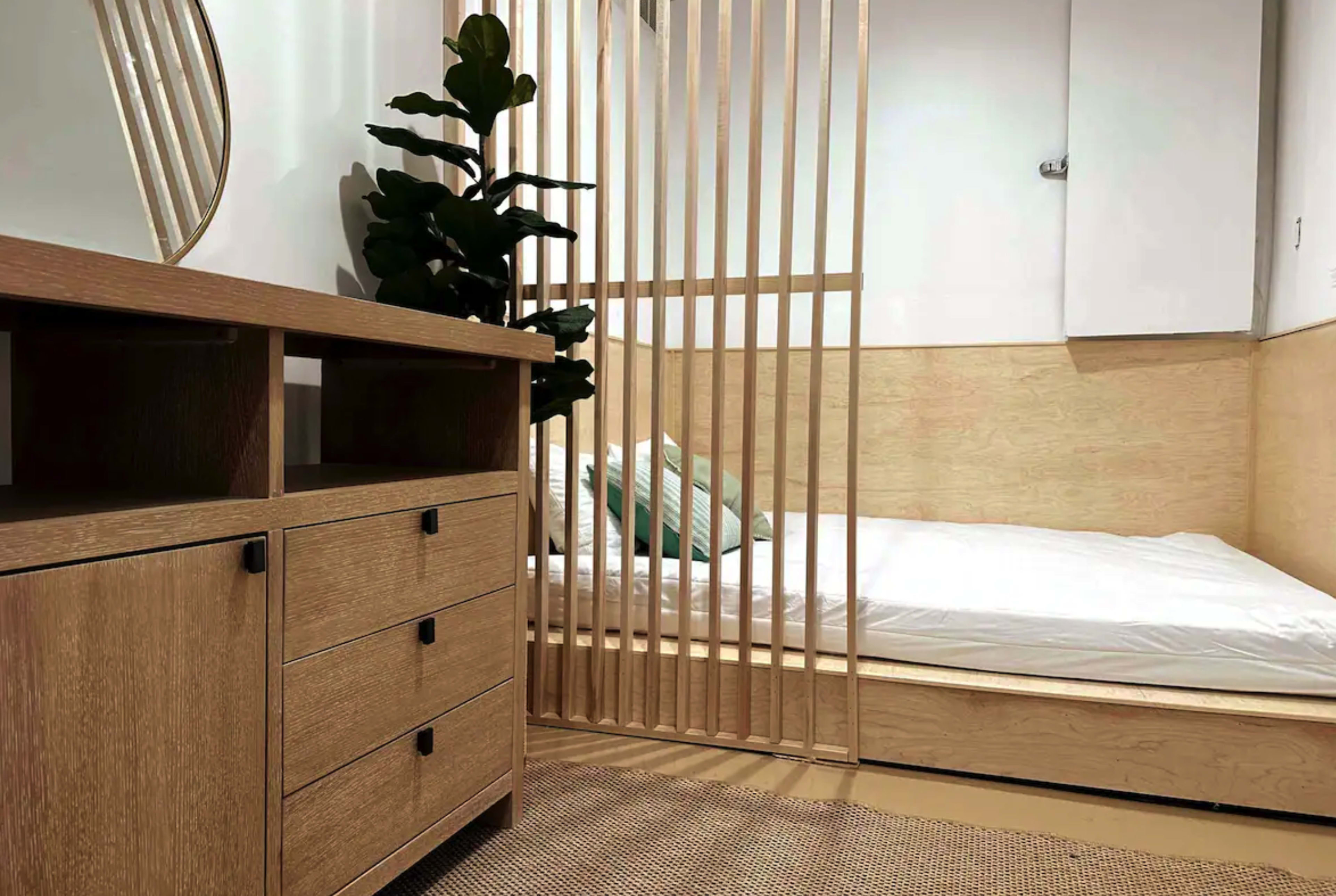 A minimalist bedroom design featuring a bed with sparse bedding, a wooden dresser with drawers, and a indoor plant separated by vertical wooden slats.