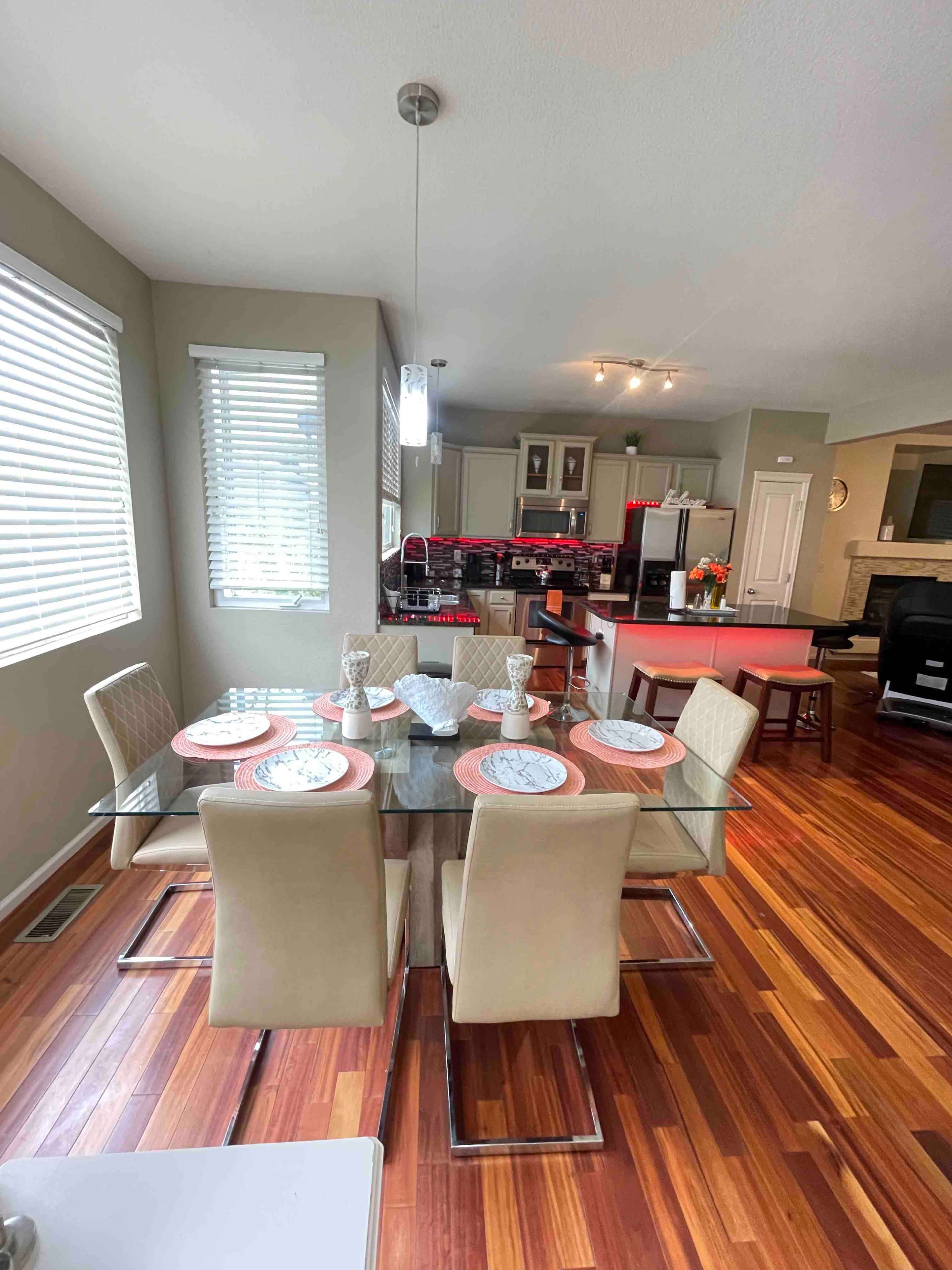 A modern dining area features a glass table set for four, with a wooden floor and an open kitchen in the background.