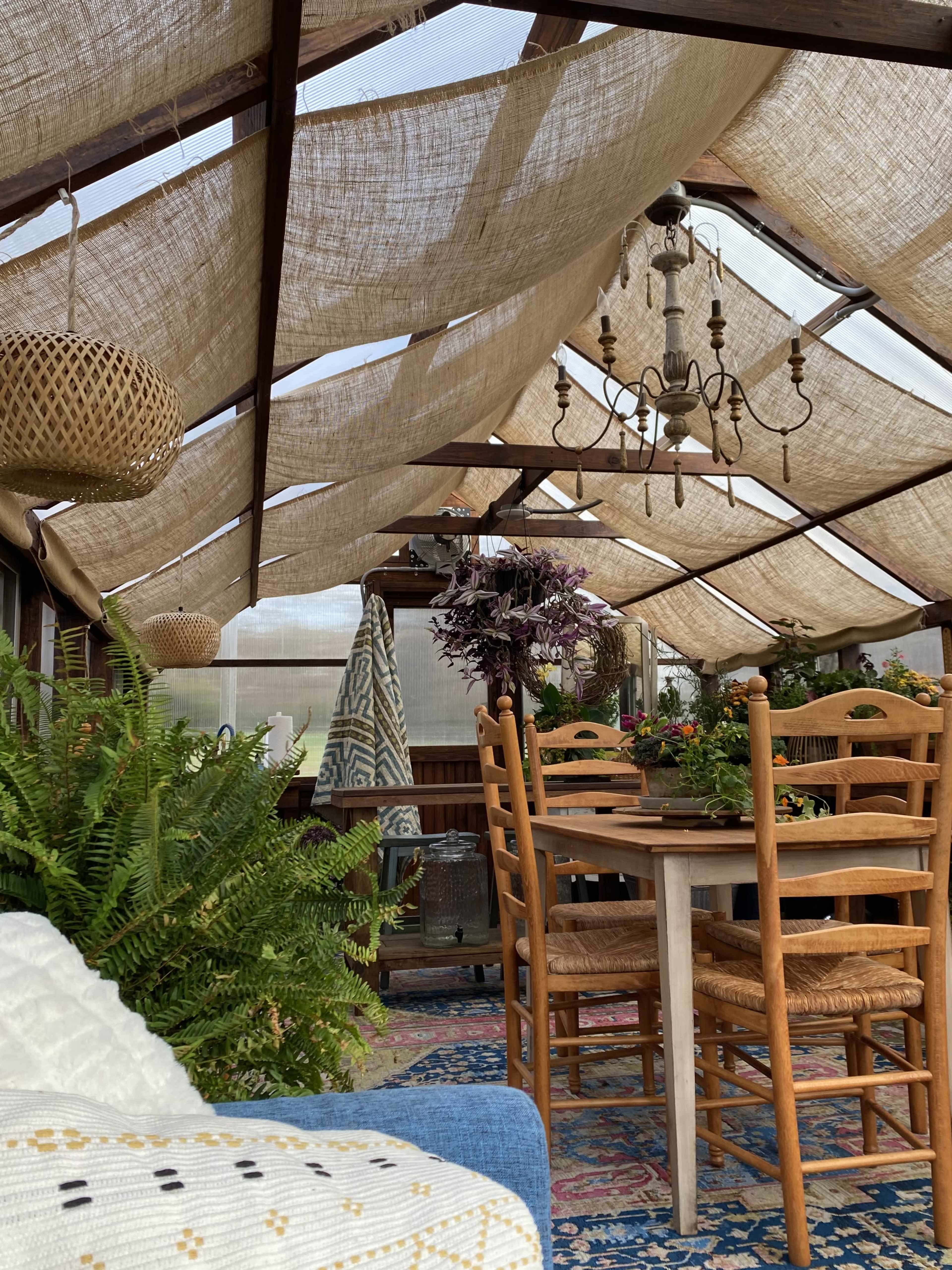 The image shows a sunlit greenhouse with a dining table surrounded by chairs, decorated with plants and hanging fixtures.