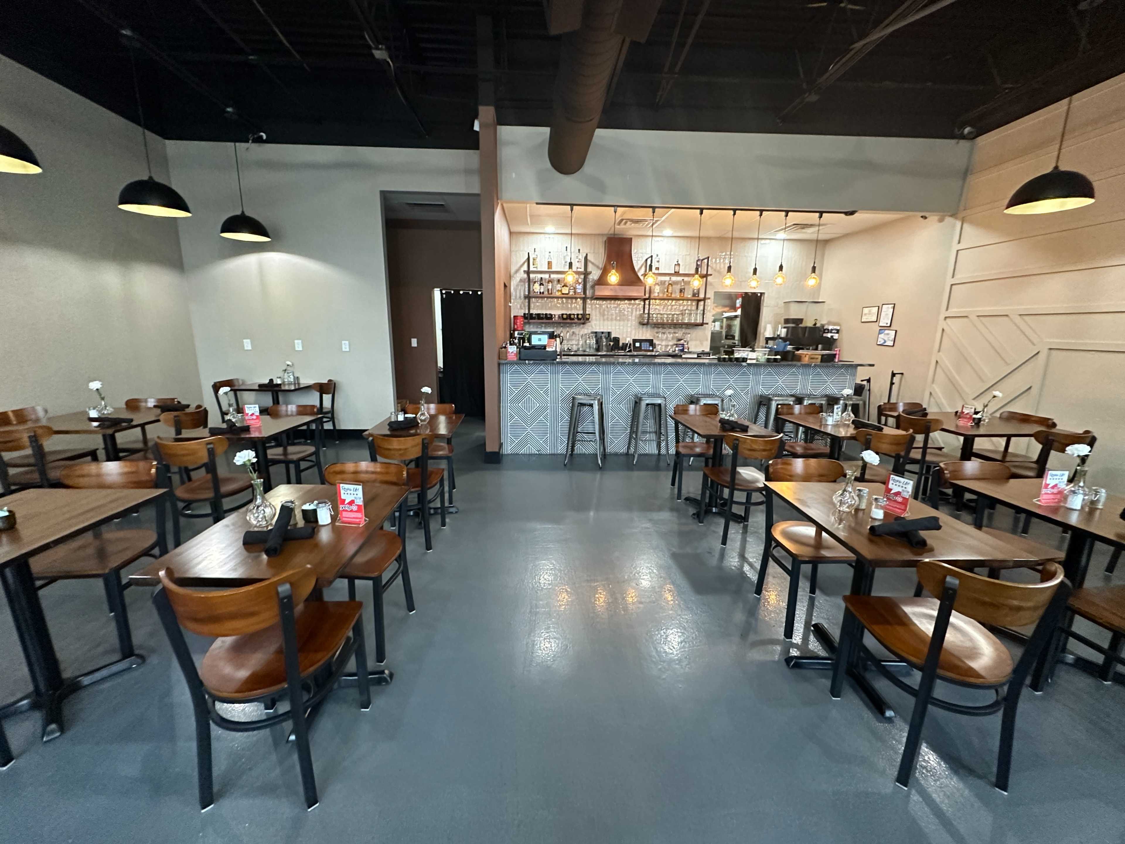 A spacious restaurant interior with tables and chairs arranged in a symmetrical layout, facing a bar area with a counter and various decorations.