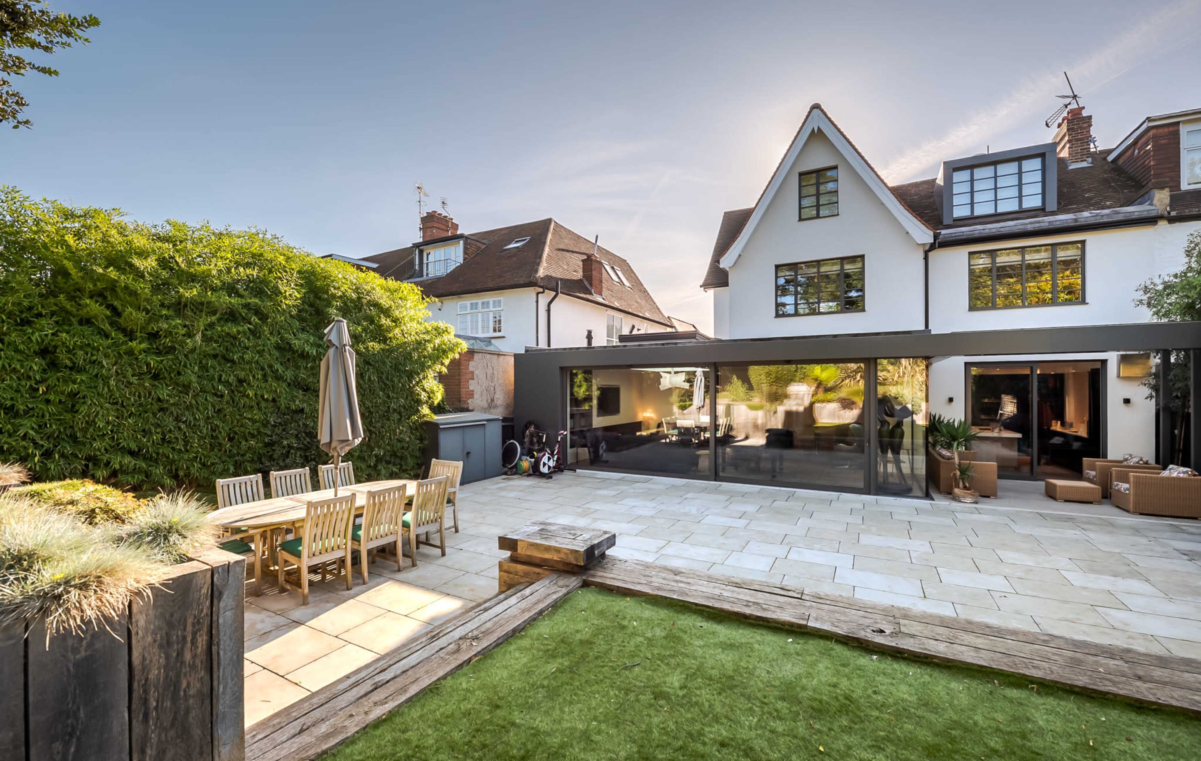 The image shows a modern outdoor patio with a dining table and chairs, lush greenery, and a large glass wall that opens up to a contemporary home.