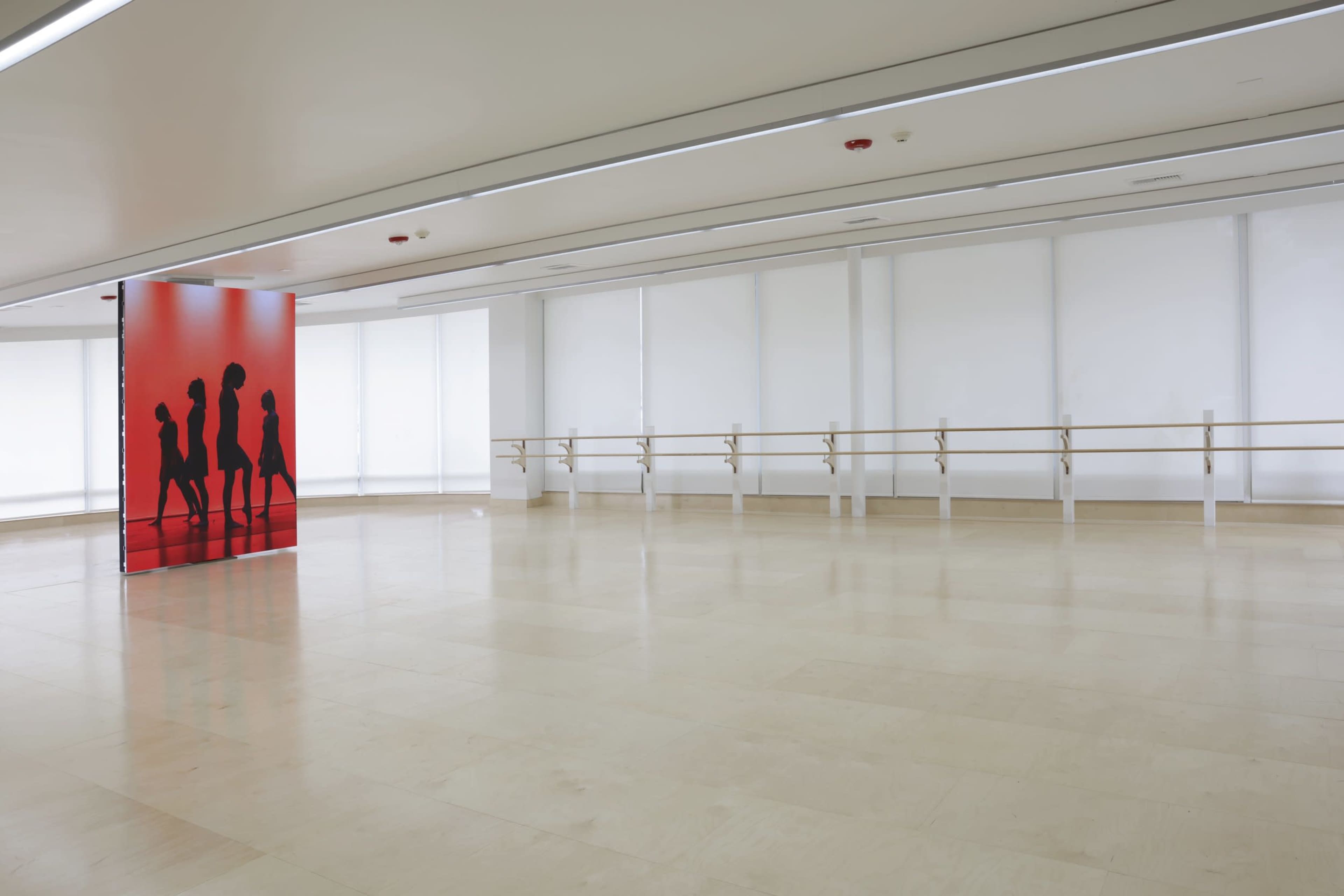 An empty dance studio with a mirrored wall, wooden ballet barres, and a large red panel featuring silhouettes of dancers.