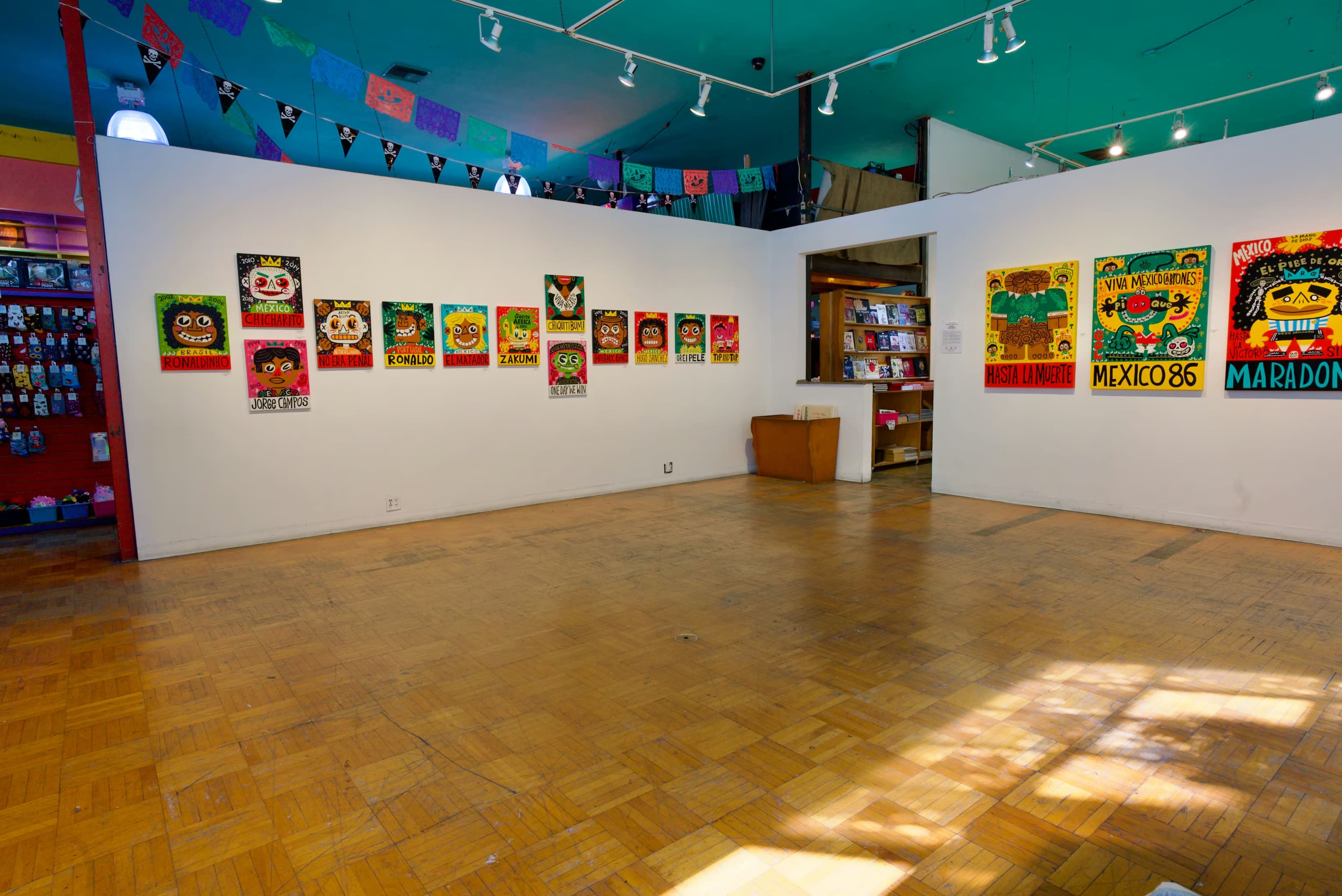 The image shows an art gallery featuring colorful posters on white walls, with wooden flooring and shelves displaying various items.