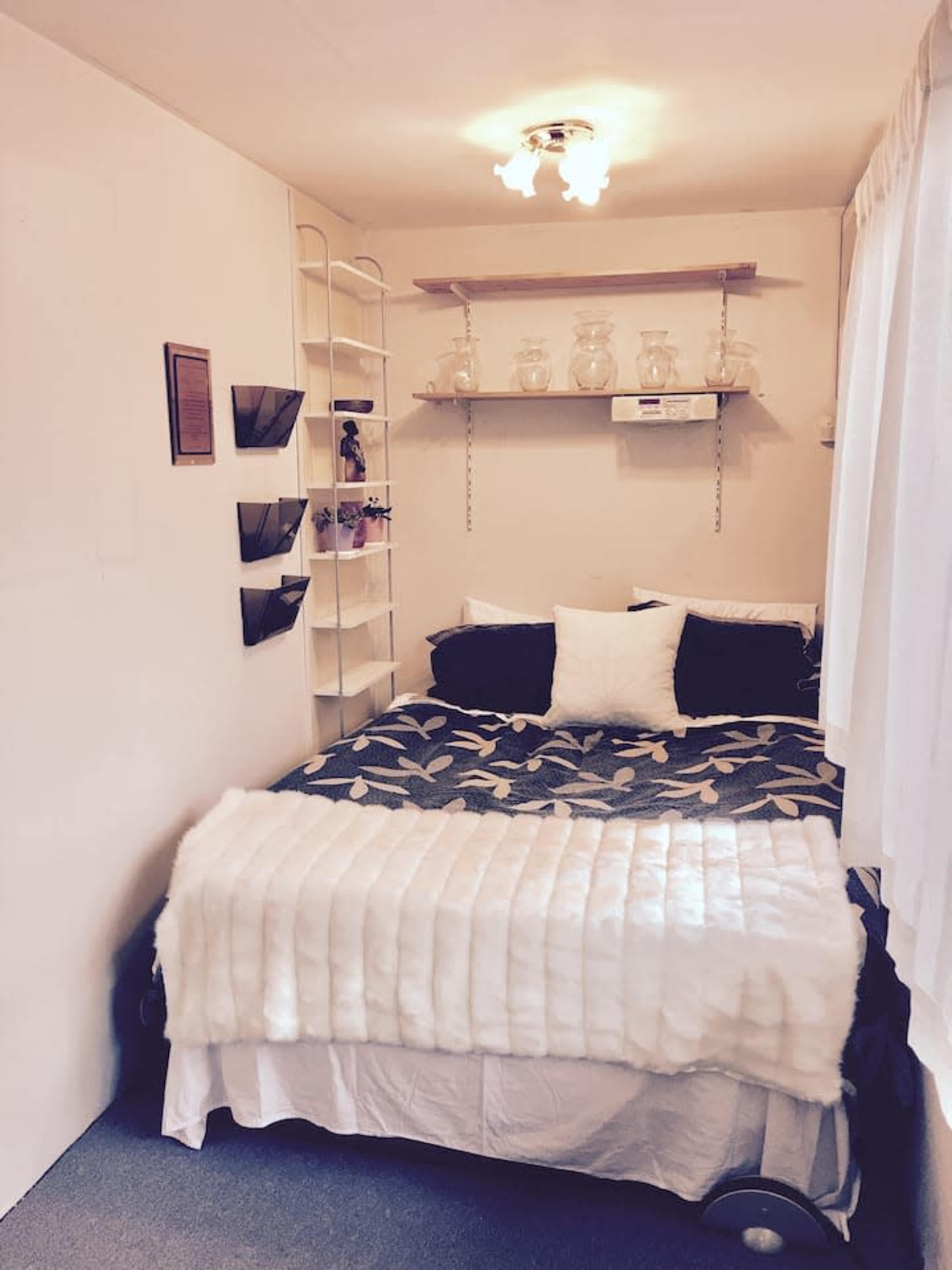 The image shows a small bedroom featuring a bed with a blanket, pillows, shelves with jars, and a wall-mounted storage unit.