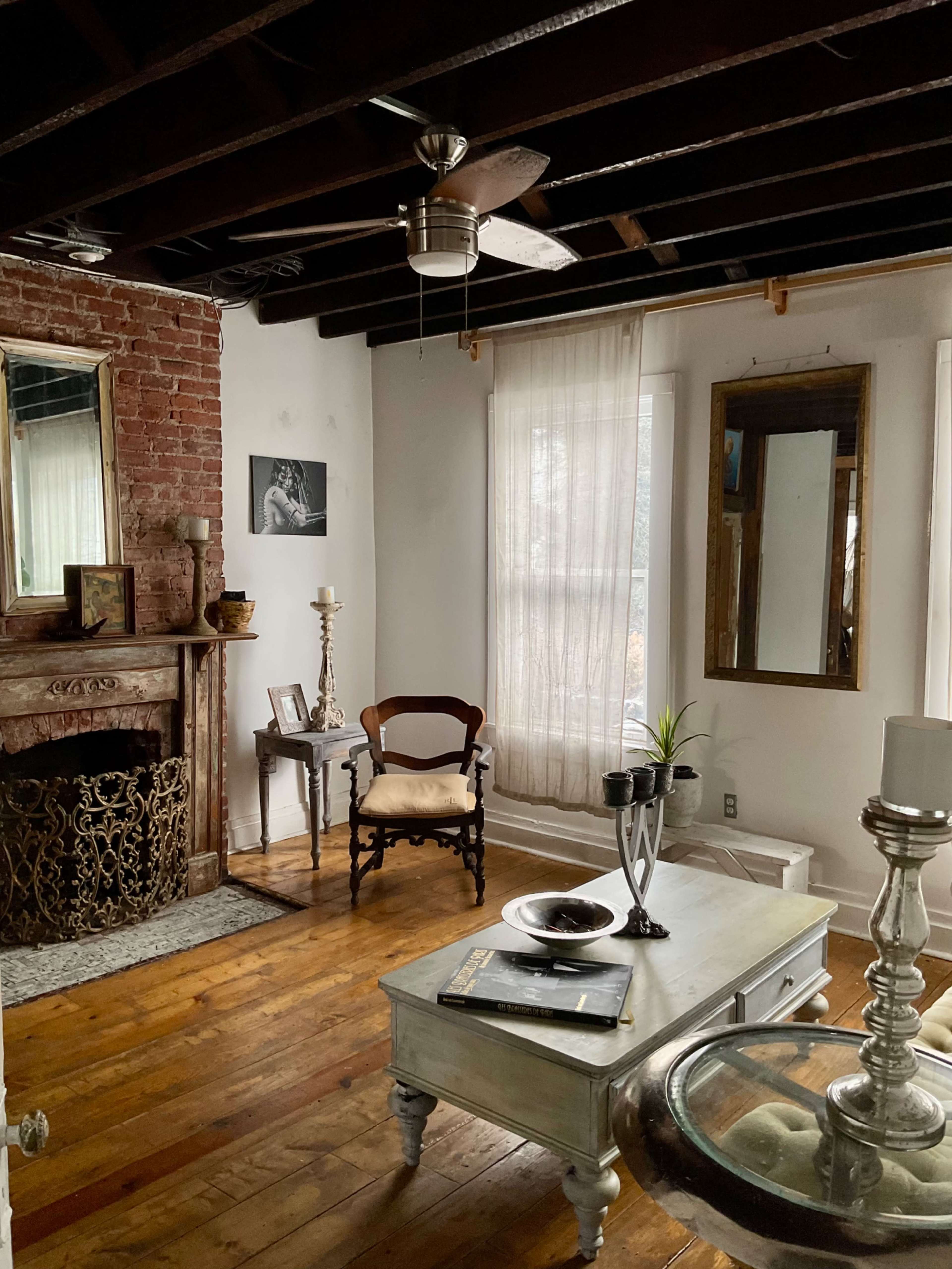 A cozy living room featuring a brick fireplace, a wooden floor, a vintage mirror, and natural light filtering through sheer curtains.