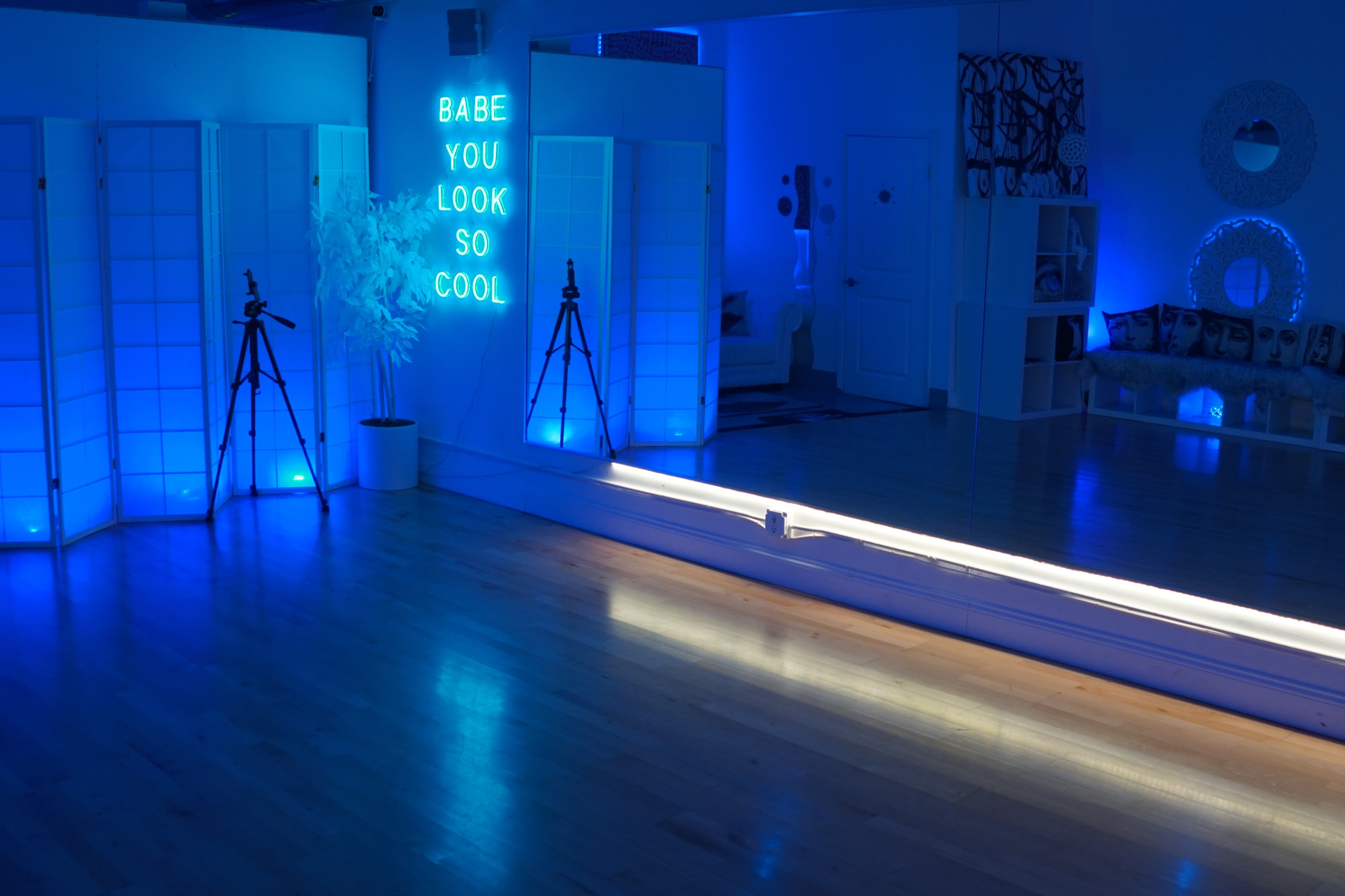 A dimly lit studio with blue lighting, featuring a mirrored wall, two tripods, and a neon sign that reads "Babe, you look so cool."